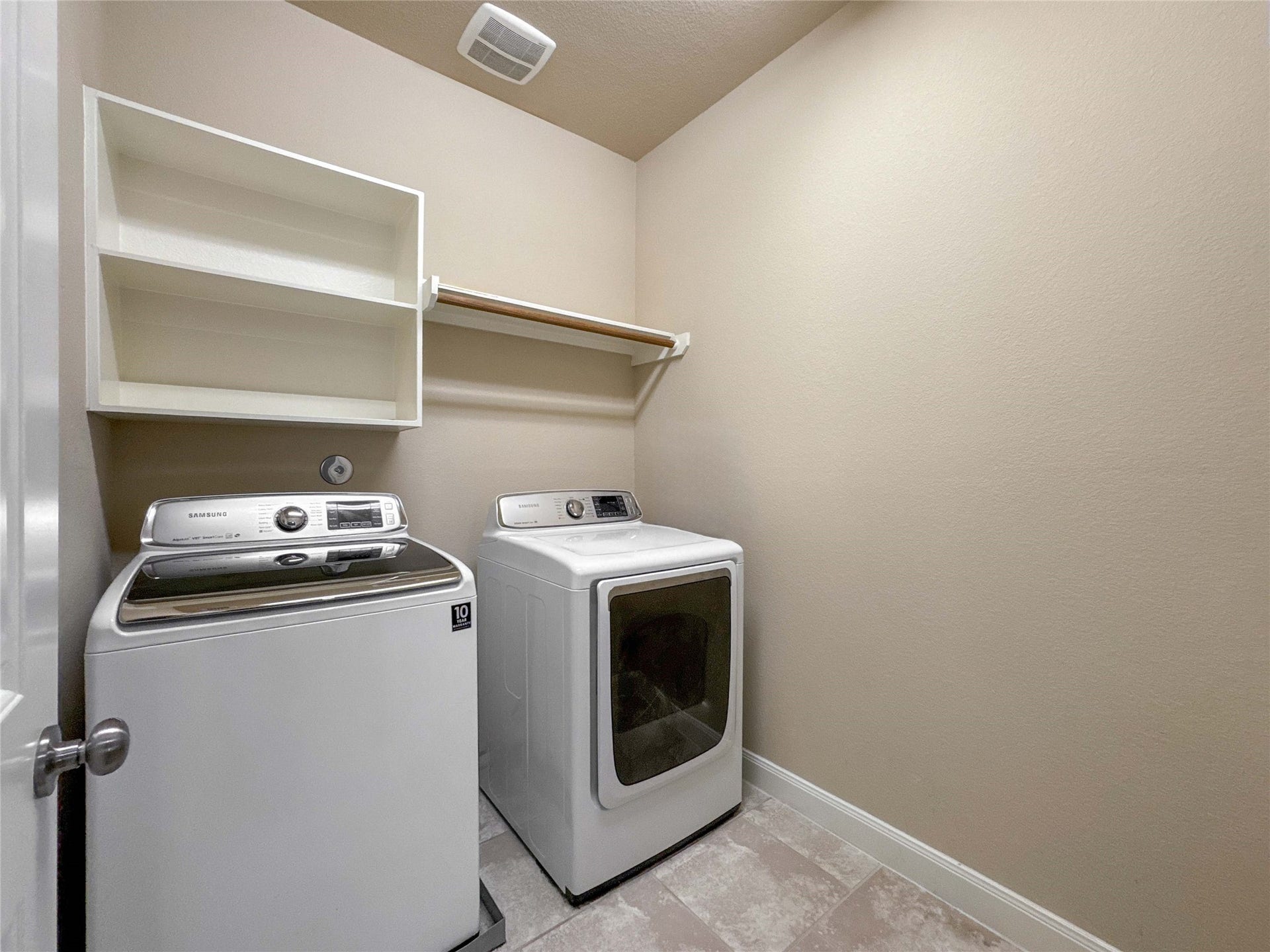 This photo shows a compact laundry room with a washer and dryer, a shelf for storage, and neutral-colored walls and flooring.