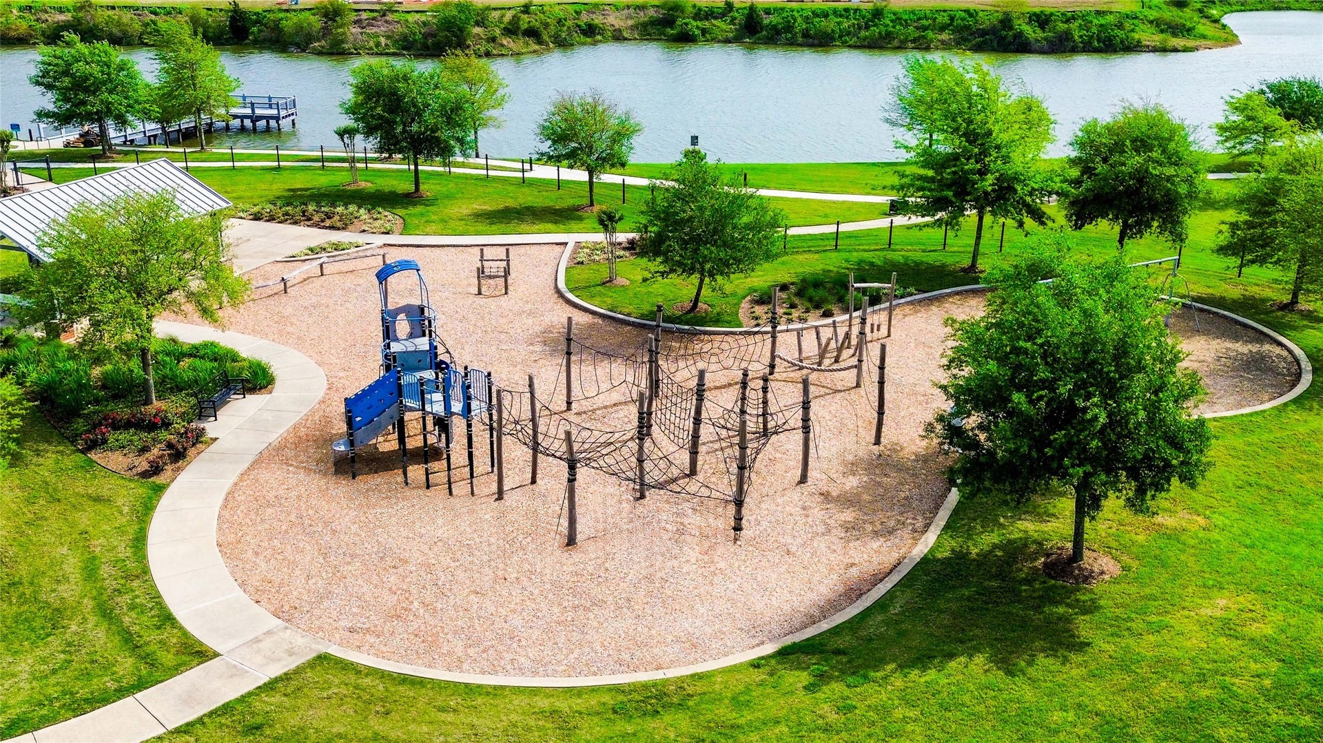 This photo showcases a scenic playground next to a lake, featuring a climbing structure, slide, and a picnic area. Surrounded by lush greenery and walking paths, it's an ideal spot for family activities and relaxation.