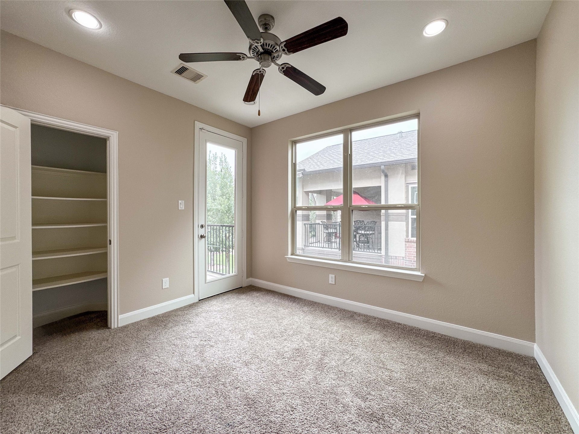This secondary bedroom located on the second floor room features plush carpeting, a ceiling fan, and neutral walls. Natural light streams through a large window and a glass door, which opens to a small balcony. A closet with built-in shelves offers ample storage space.