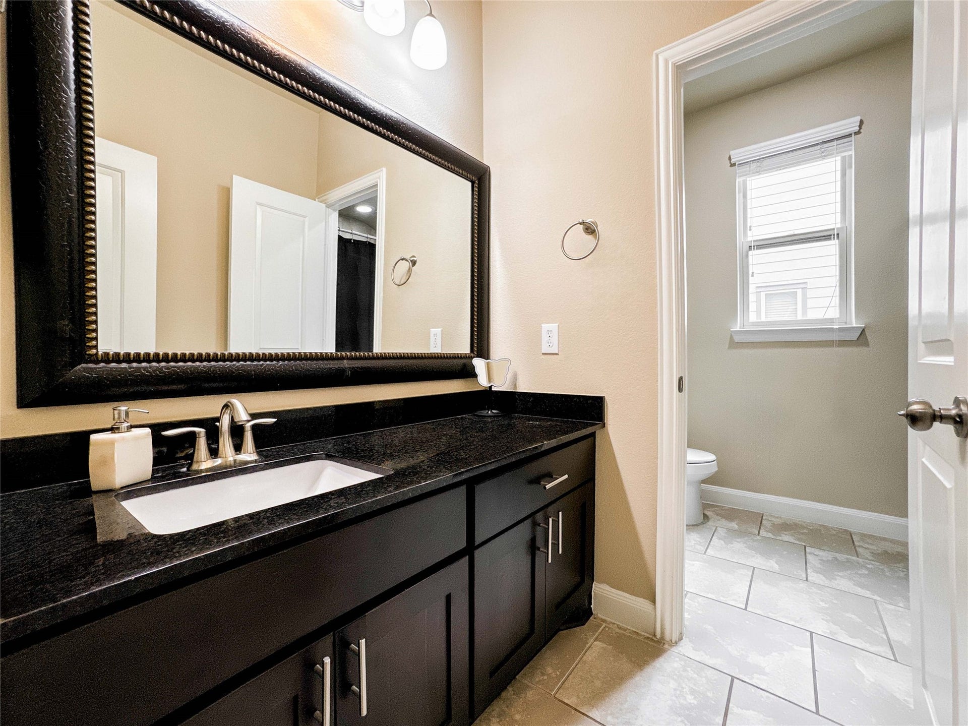 This bathroom features a sleek dark vanity with a large mirror, modern fixtures, and ample storage. The tiled floor leads to a separate toilet area with natural light from a window.