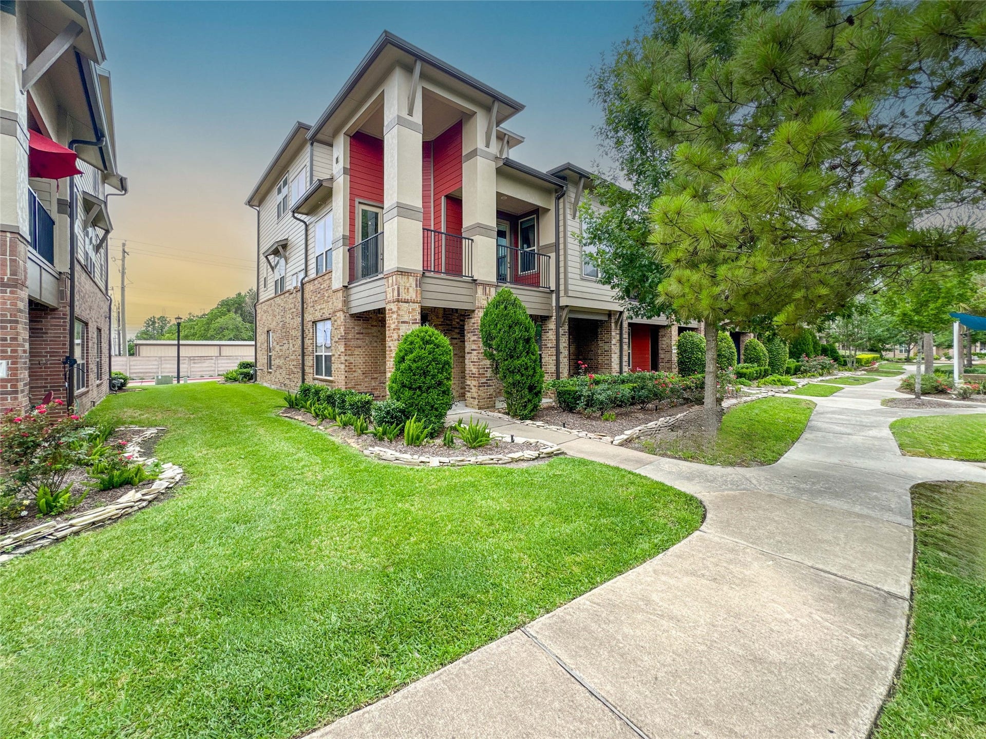 Modern two-story townhouse with a mix of brick and siding exterior, featuring a covered balcony. Well-maintained landscaping with lush green lawns and shrubs. Paved pathways lead through the community, creating a welcoming ambiance.