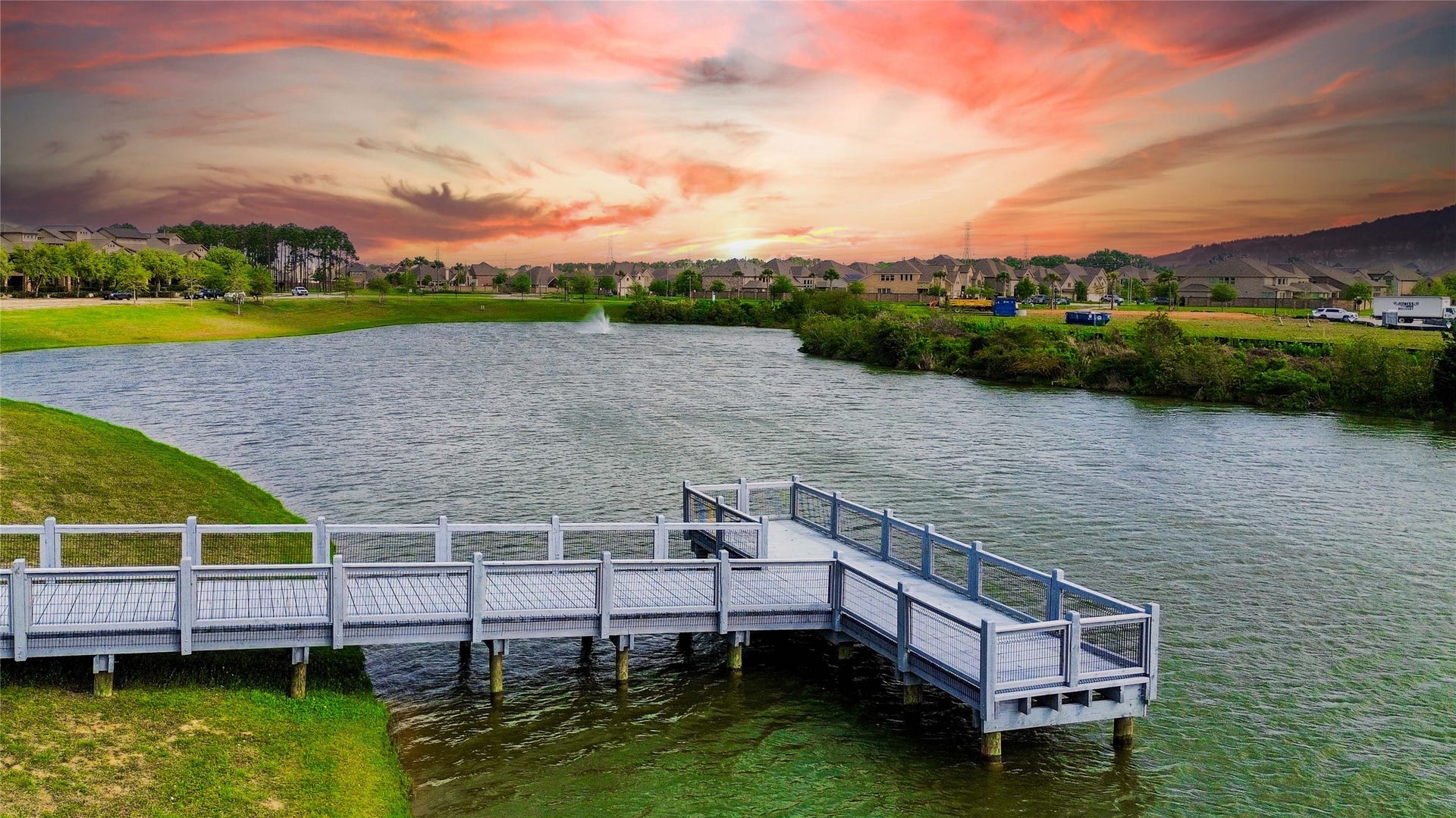 This photo showcases a serene lakeside view with a wooden dock extending over the water, set against a vibrant sunset. The surrounding area features well-maintained greenery and a nearby residential community, offering a peaceful and picturesque environment.