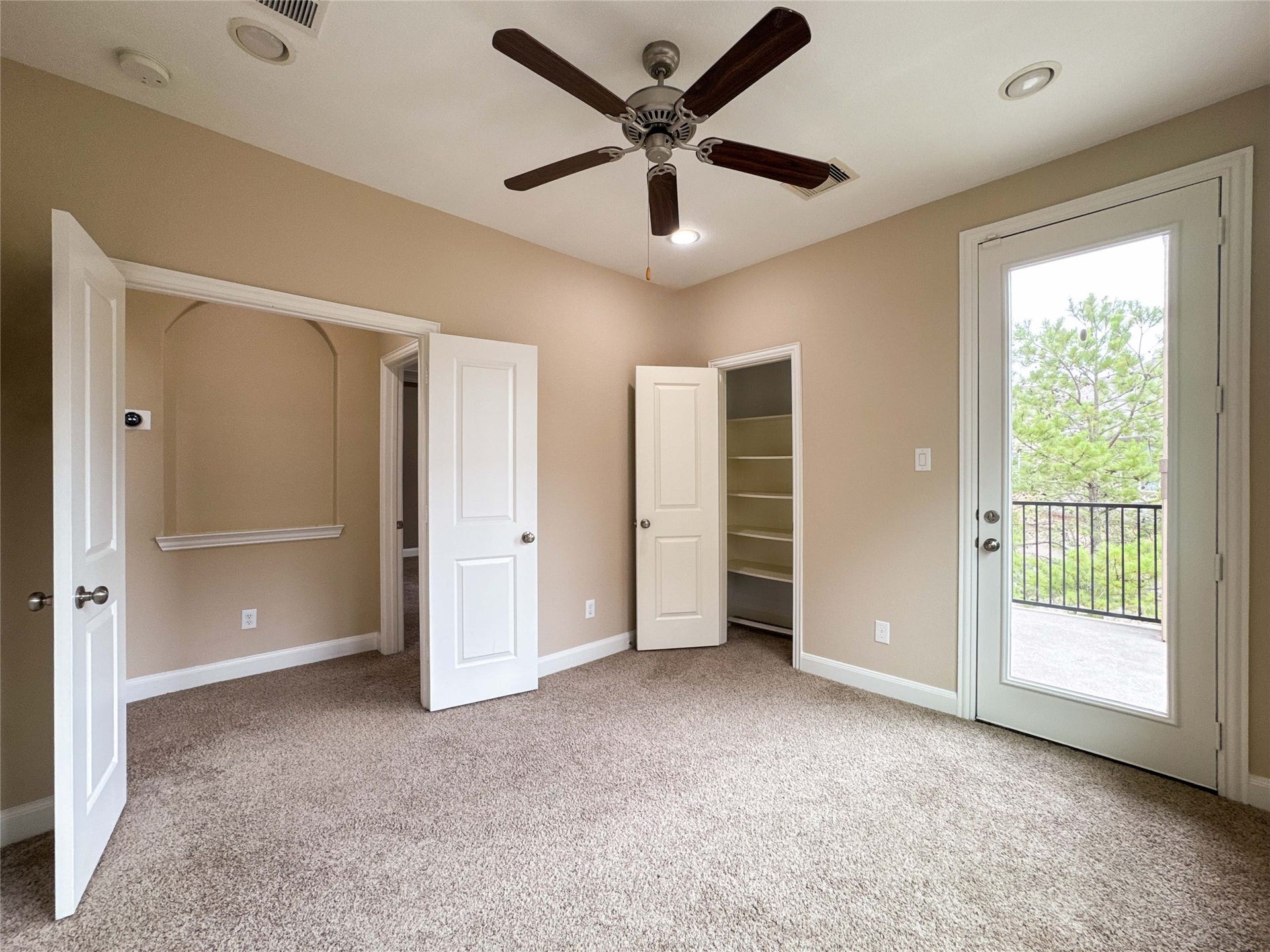 Alternate view of the room features neutral walls, plush carpet, and a ceiling fan. It includes a closet, a recessed shelf, and French doors leading to a private balcony with a view of trees.