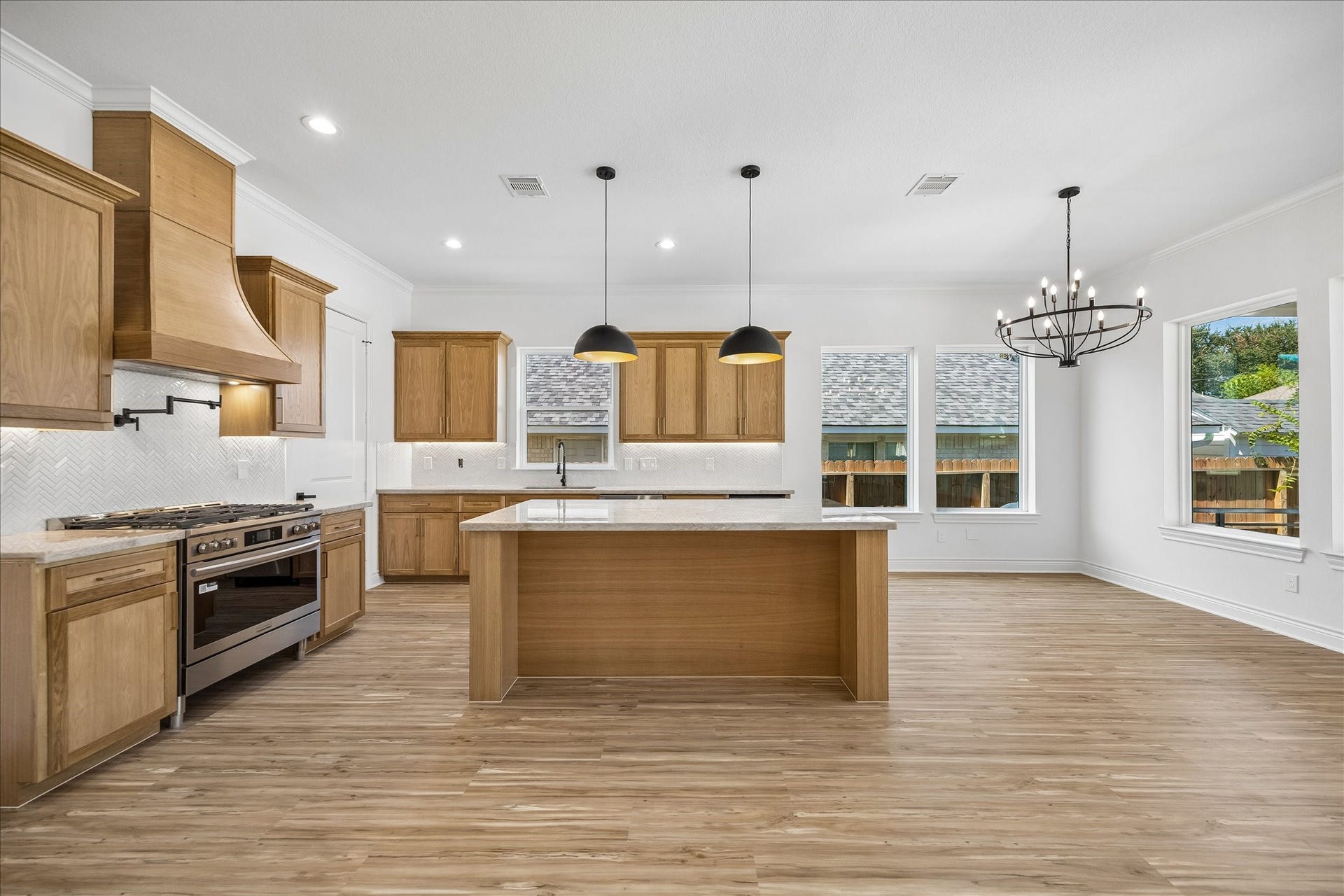 Beautifully appointed kitchen featuring custom white oak cabinetry, a spacious center island, elegant herringbone tile backsplash, stainless steel appliances, and stylish pendant lighting, all complemented by warm wood-look flooring and an open dining area.