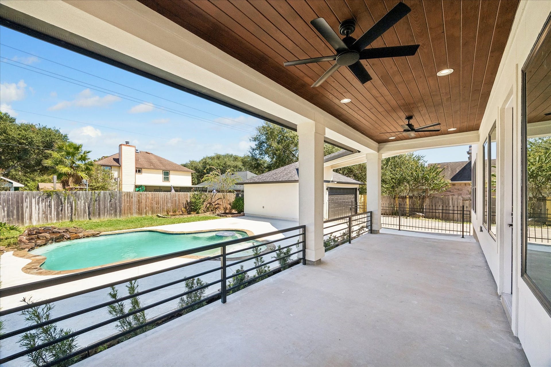 A covered outdoor patio with a wood planked ceiling, an outdoor fan and modern wrought iron railing that over looks the pool.