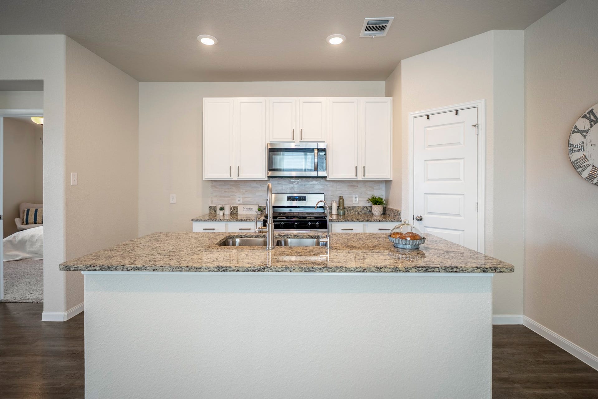 2355 Mount Locke Path, Conroe, TX 77301 Standing at the island, this view shows how the kitchen sits right between the homes main gathering spaces and the first-floor primary suite. The doorway on the left leads into the primary bedroom, while the pantry is tucked behind the door to the right. Granite counters provide plenty of prep space around the sink, with the upgraded backsplash, cabinet hardware and stainless appliances all in view.