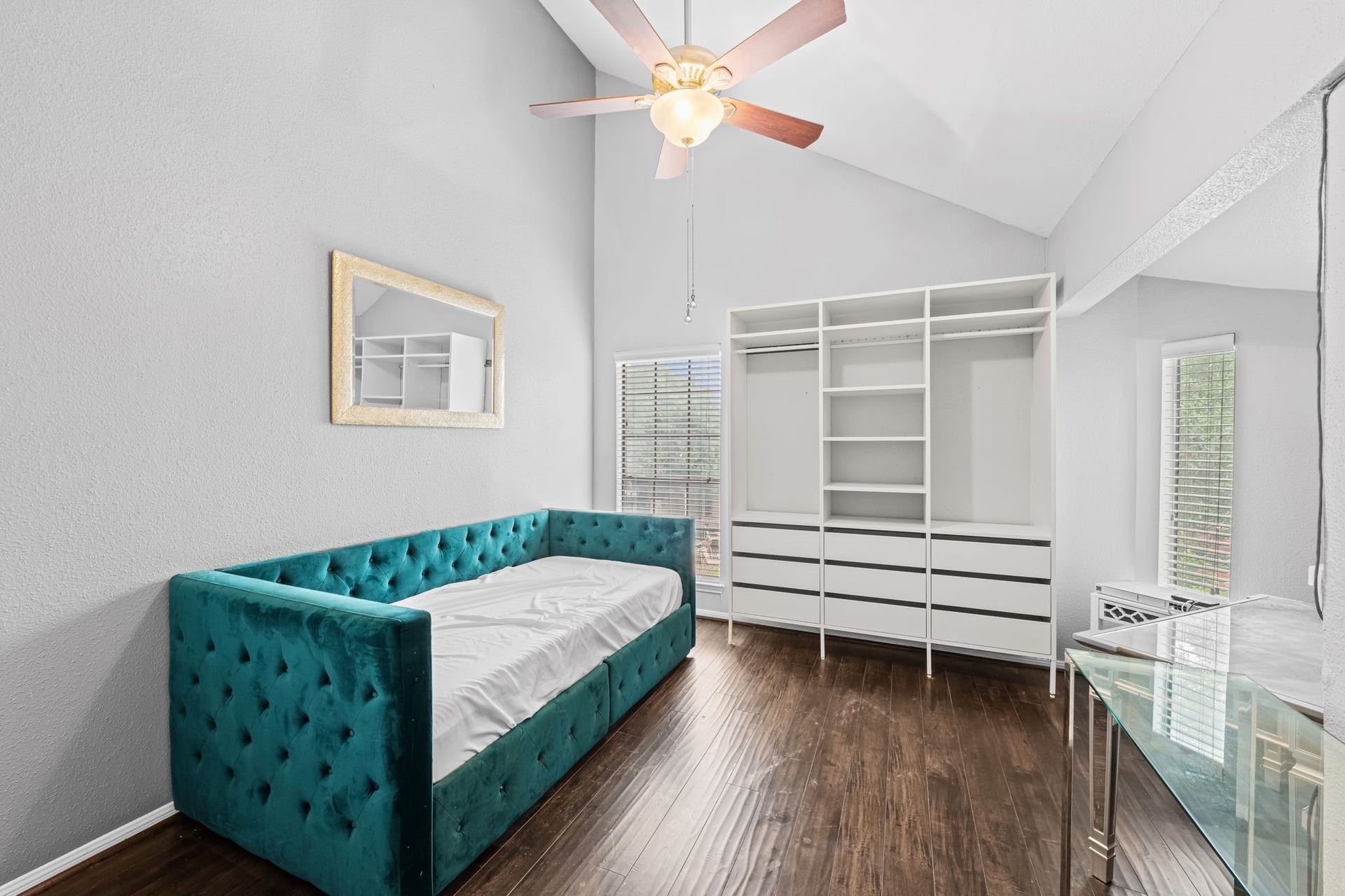2211 S Kirkwood Road, #1, Houston, TX 77077 The secondary bedroom has a cathedral-height vaulted ceiling that rises well above what any standard second-floor room would offer. A ceiling fan sits at the apex, two windows on adjacent walls bring natural light from both directions, and a full-wall built-in shelving and wardrobe system in white provides substantial organized storage without consuming floor space. It's a room that earns its footprint.
