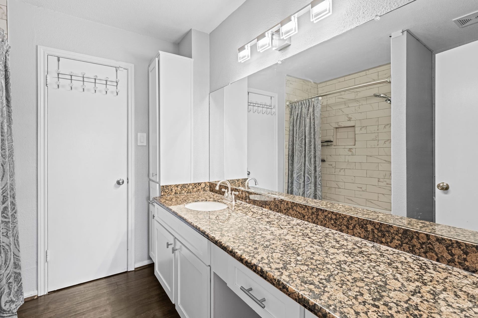 2211 S Kirkwood Road, #1, Houston, TX 77077 The primary bath is anchored by a wide dark speckled granite vanity that runs nearly the full width of the wall substantially more counter surface than a standard condo bath delivers. White shaker cabinetry, a full-width mirror, and a modern rectangular light bar complete the vanity wall, with white subway tile in the shower visible in the mirror's reflection.