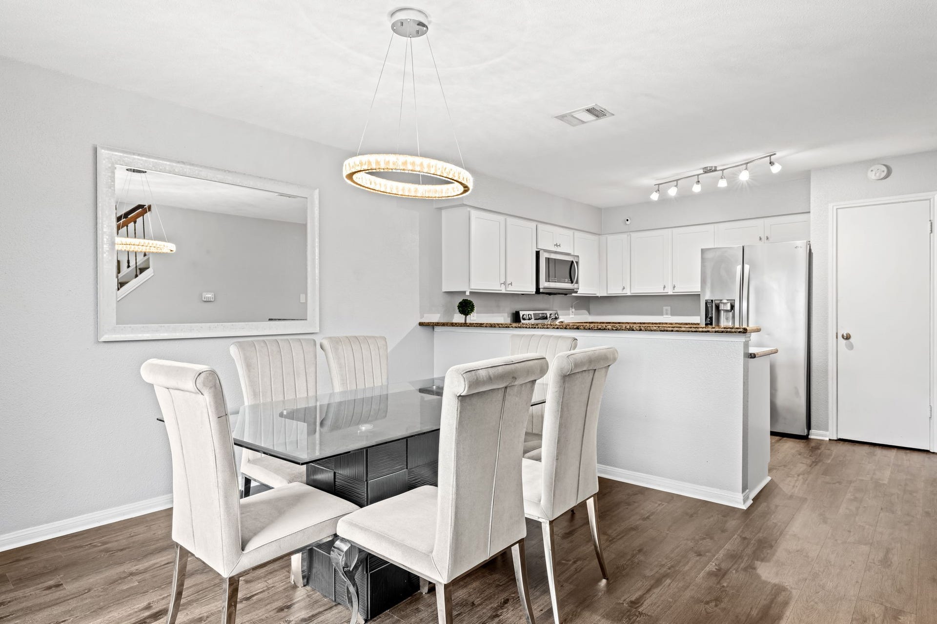 2211 S Kirkwood Road, #1, Houston, TX 77077 The dining area and kitchen read as one connected space from this vantage the crystal ring pendant chandelier hangs above the dining area while the breakfast bar peninsula, white shaker upper cabinetry, and stainless steel refrigerator are all visible through the opening beyond. It's a layout that keeps the cook engaged with the room rather than separated from it.