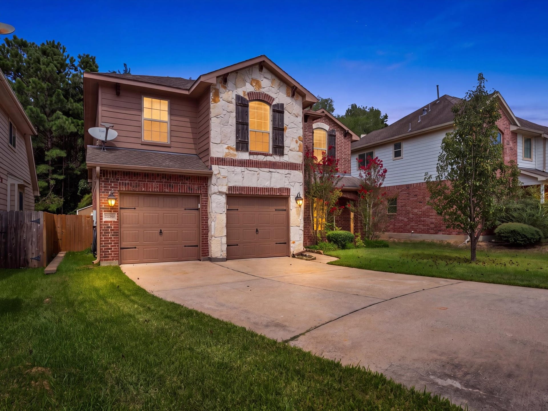 Stunning twilight view highlighting the impressive stone and brick exterior, rustic shutters, and cozy warm interior lighting. Features a spacious concrete driveway, attached two-car garage, and charming front landscaping.