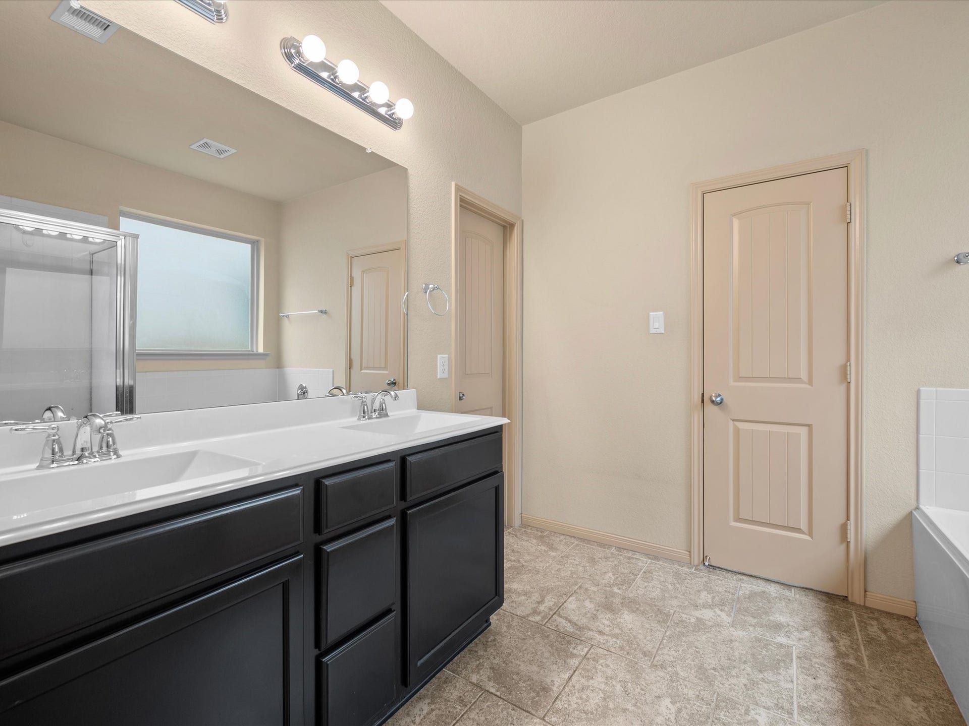 Primary en-suite bathroom boasting a double vanity with dark cabinetry, wide vanity mirror, tile flooring, walk-in shower, and dedicated closet space.