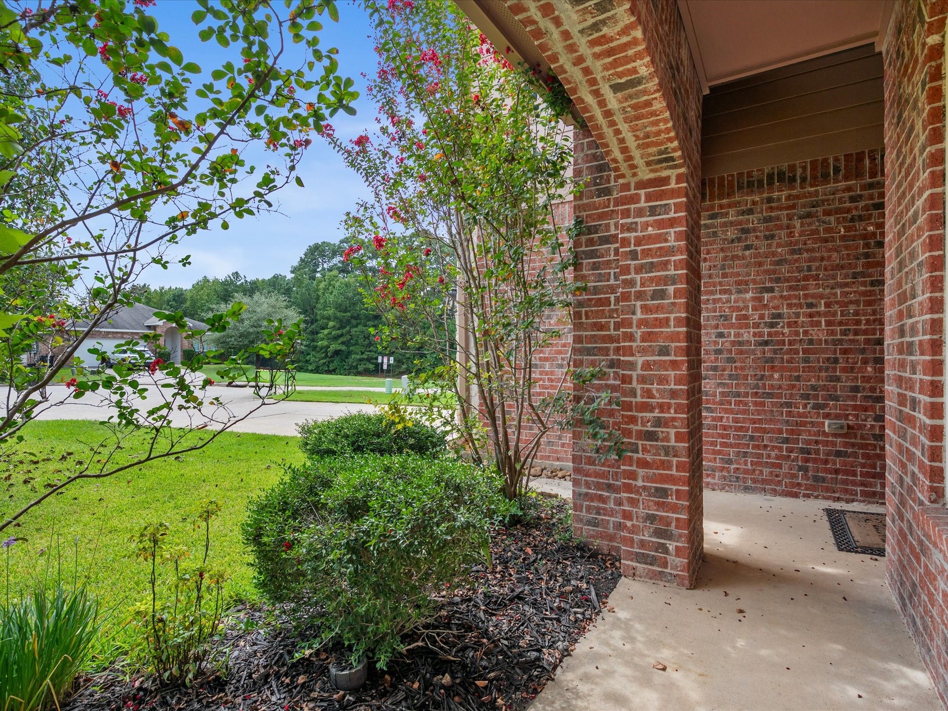 View looking out from the covered front porch framed by a classic brick archway. Highlights the peaceful neighborhood surroundings, manicured front garden beds with dark mulch, and lush flowering landscaping.