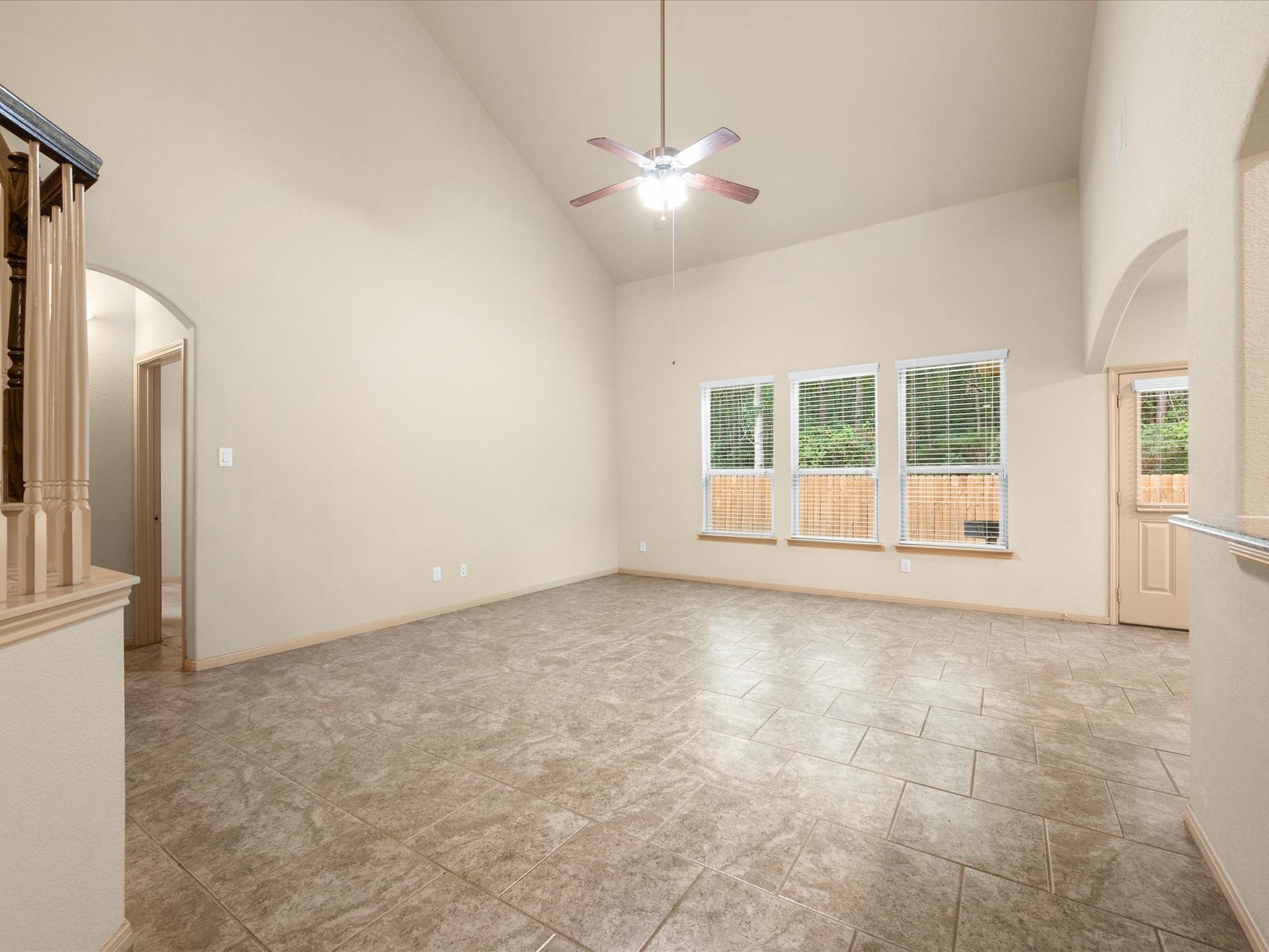 Spacious family room featuring soaring vaulted ceilings, a central ceiling fan, and durable tile flooring throughout. A wall of large windows floods the space with natural light while offering views of the private backyard.