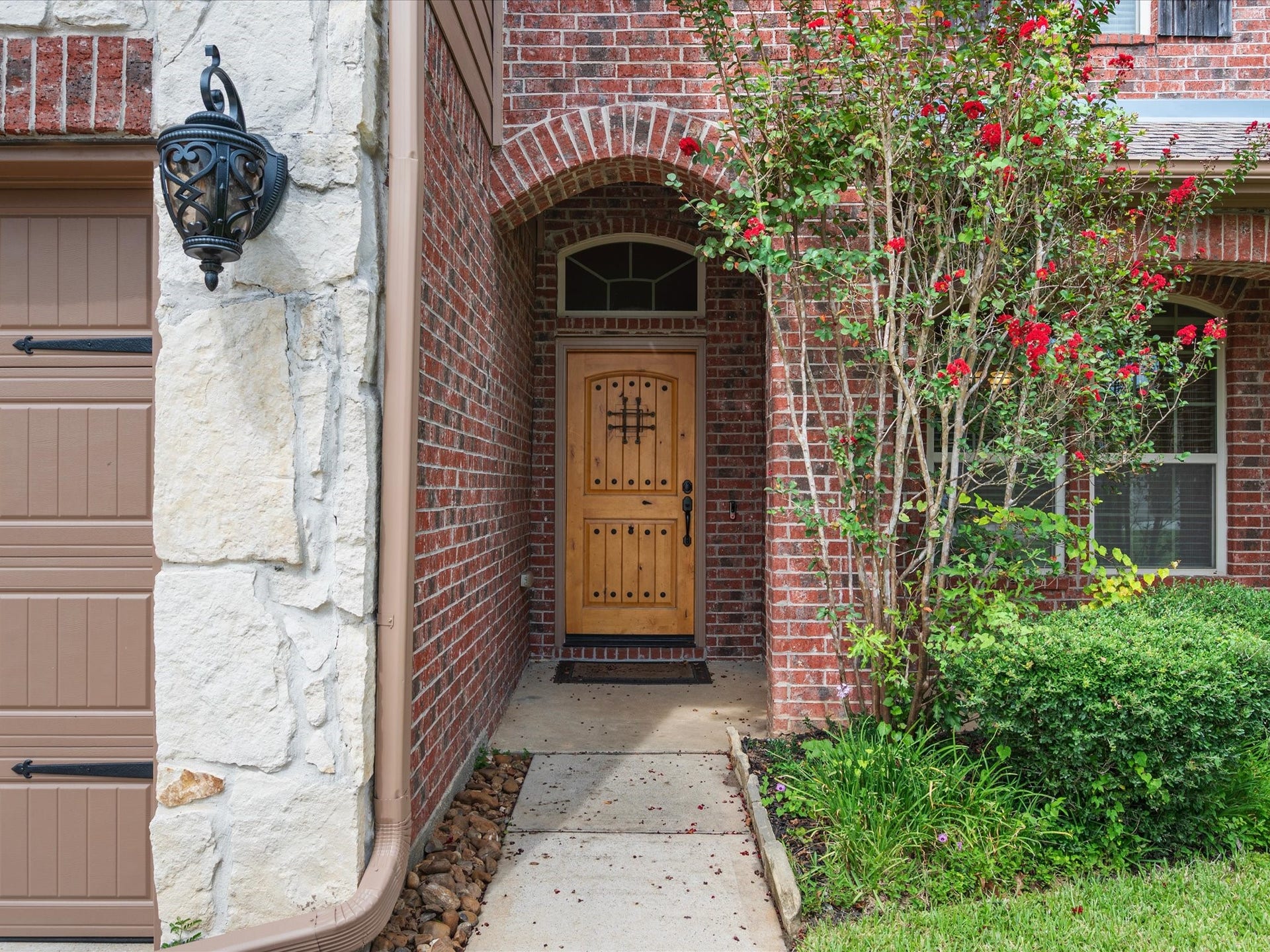 Welcoming covered entryway featuring a rustic wooden front door with ornate iron grill work and a transom window above. Surrounded by rich brick masonry, stone accents, classic exterior lantern lighting, and lush flowering landscaping.