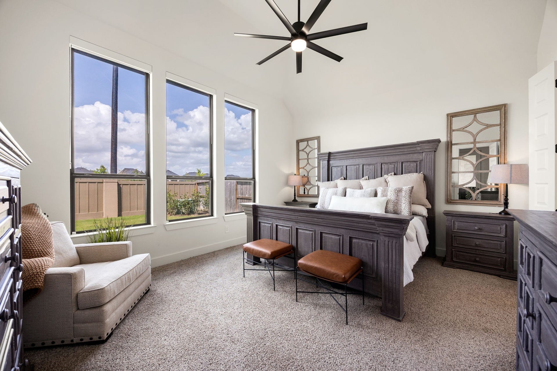 31322 Crescent Hill Drive, Fulshear, TX 77441 A serene primary retreat. Three towering, black-trimmed vertical windows with pull down blackout shades flood this spacious bedroom with natural light, showcasing plush carpeting, soaring vaulted ceilings, and a modern multi-blade ceiling fan with integrated lighting.