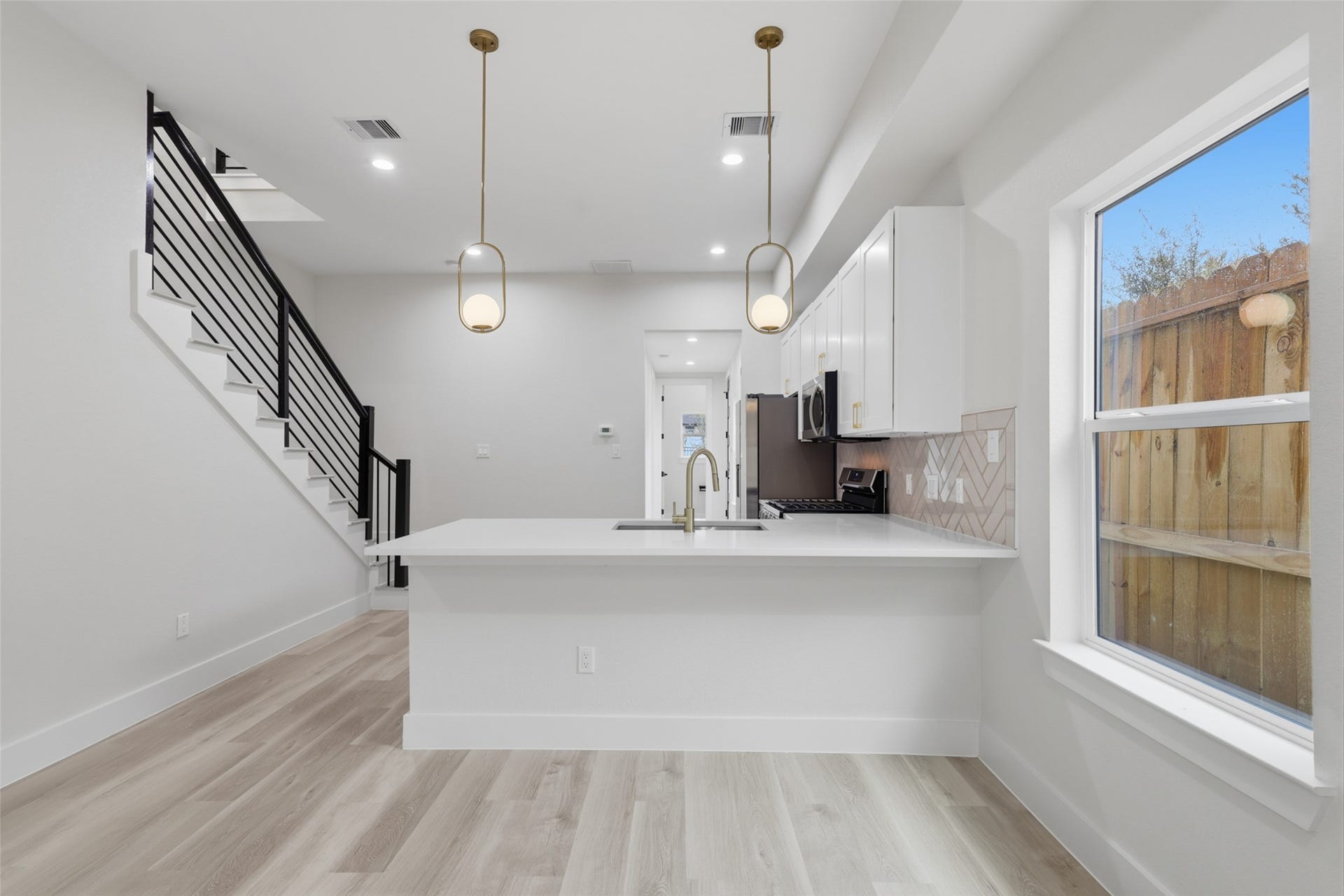 3205 Berry Street, #A/B, Houston, TX 77004 Modern kitchen with sleek white cabinetry, stainless steel appliances, and stylish pendant lighting. The open layout flows into a bright space with a wooden staircase and large window, enhancing natural light.