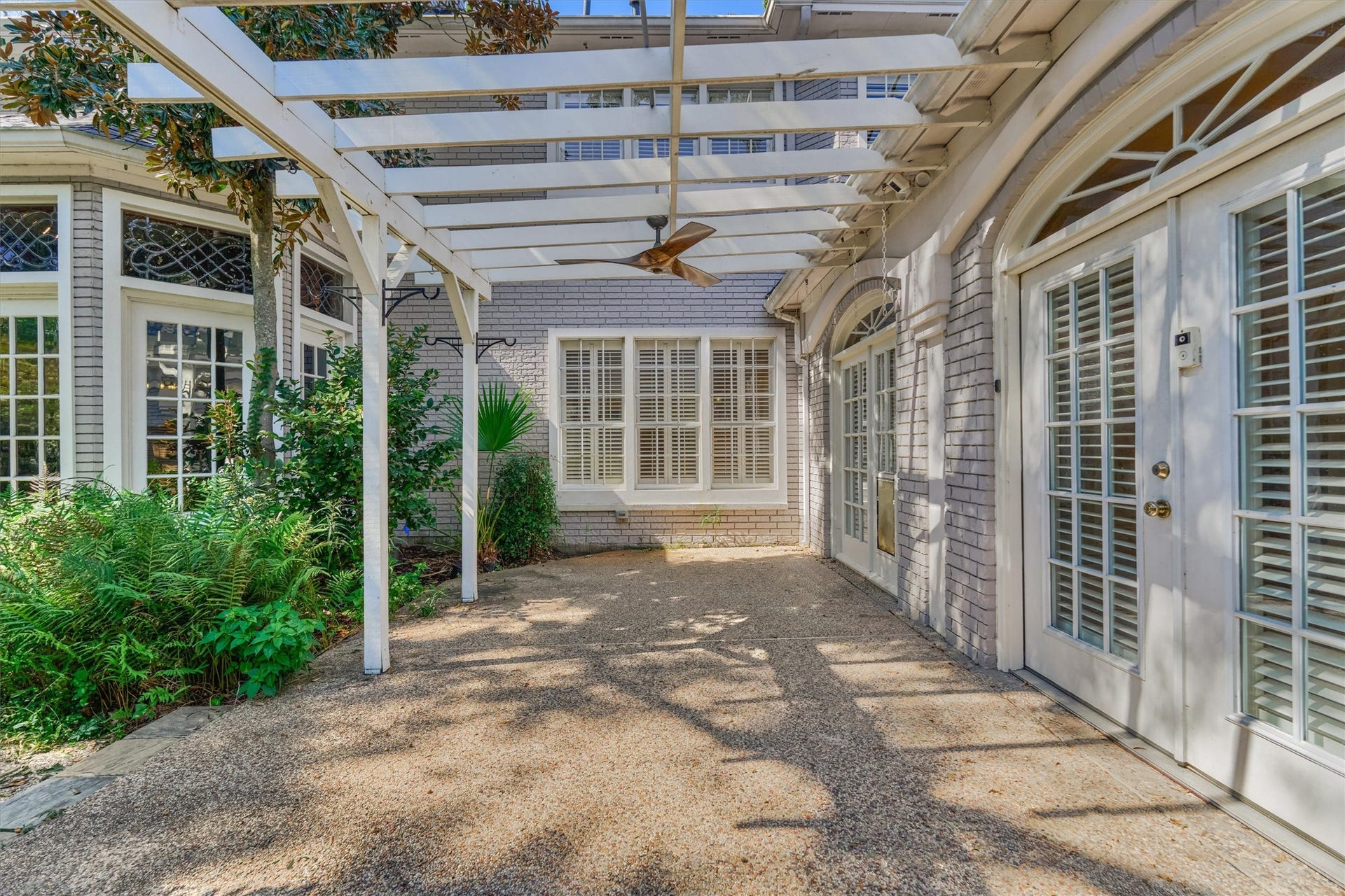 26 S Brokenfern Drive, The Woodlands, TX 77380 This outdoor patio sits off the sunroom, with a white pergola overhead and a ceiling fan below it. The aggregate floor is framed by brick walls and French doors with plantation shutters and arched transoms, and the librarys leaded glass windows are visible across the courtyard. Mature magnolia, ferns, and a palm soften the edges, and plant hooks on the pergola posts are ready for hanging baskets.