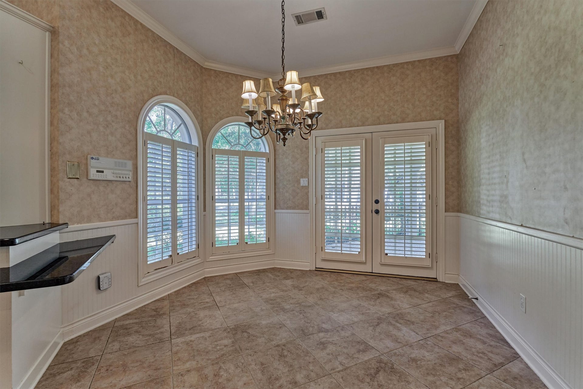 26 S Brokenfern Drive, The Woodlands, TX 77380 This breakfast area is lit by two arched windows with half-round transoms and plantation shutters, set in a gently angled bay. Beadboard wainscoting with a chair rail and deep crown molding add classic detail, and a chandelier hangs over the tile floors. A granite counter breakfast bar connects the space to the kitchen, and shuttered French doors open to the front covered patio.