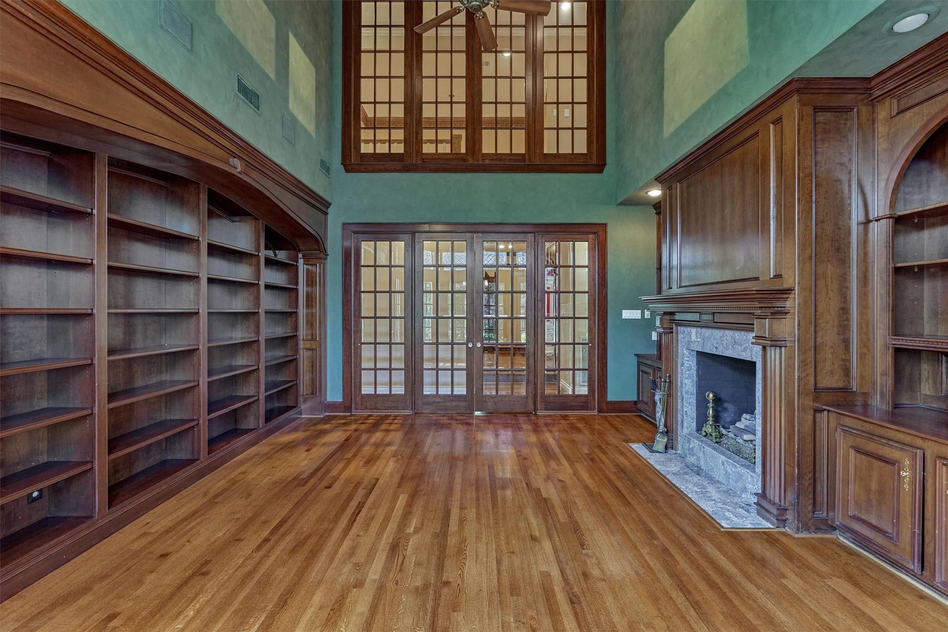 26 S Brokenfern Drive, The Woodlands, TX 77380 Another view of this two-story library shows its 19-foot ceilings and wall of custom built-in bookshelves beneath an arched cornice. The fireplace wall features raised-panel wood paneling, a carved mantel with fluted pilasters, a granite surround, and an arched display niche. Double French doors with divided lights open to the adjoining rooms, and a high interior window brings light and views between floors.
