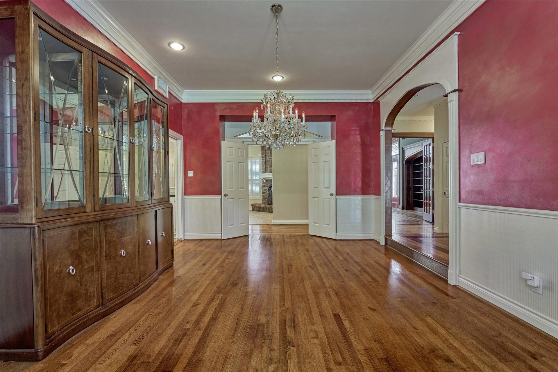 26 S Brokenfern Drive, The Woodlands, TX 77380 This formal dining room off the foyer has 10-foot ceilings, deep crown molding, and wainscoting with a chair rail. An arched opening trimmed in white leads back toward the foyer, while double doors open to the adjoining family room with its brick fireplace. A crystal chandelier and recessed lighting highlight the hardwood floors, with plenty of space for a large table and a china cabinet.