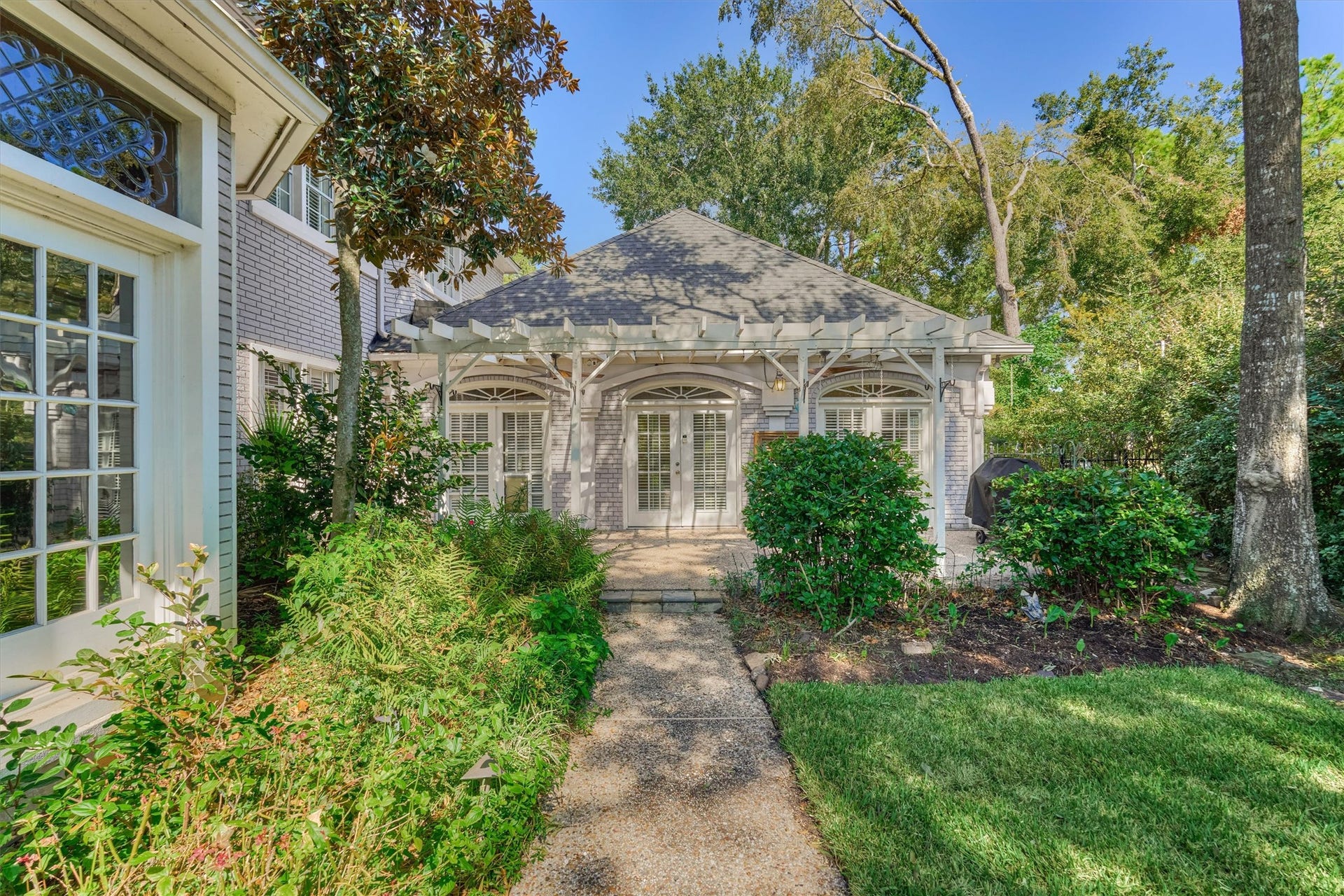 26 S Brokenfern Drive, The Woodlands, TX 77380 Another angle of the backyard patio shows how easily the house flows outdoors. Three sets of French doors open to a shaded, pergola-covered seating area with room for a grill, dining, and lounging. A winding walkway leads from the lawn to the patio, and tall trees and lush plantings keep it private and peaceful.