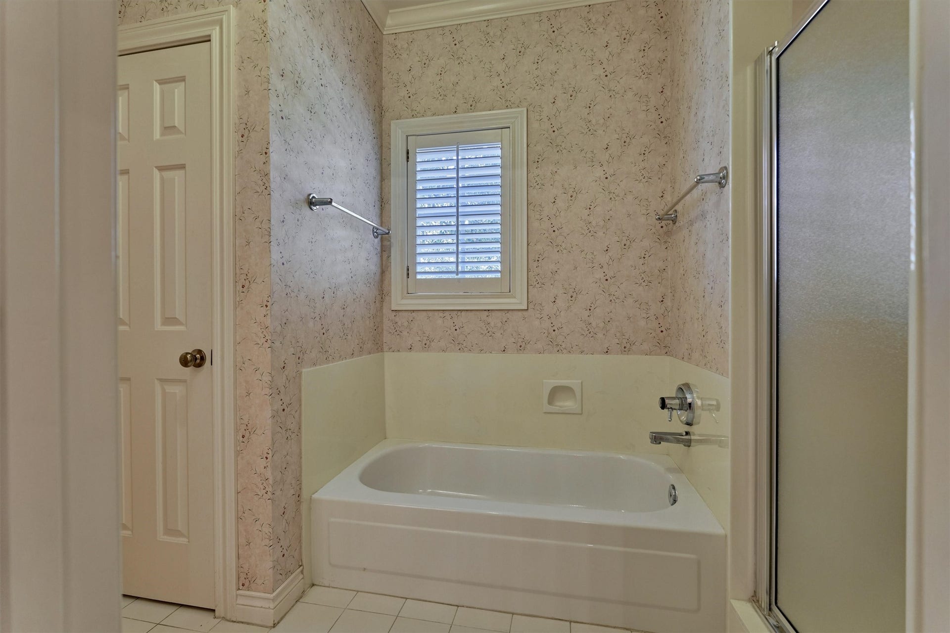 26 S Brokenfern Drive, The Woodlands, TX 77380 This shared bath has a soaking tub with a tiled-style surround, built-in soap niche, and chrome fixtures, beneath a window with plantation shutters. A separate shower with a frosted glass door sits beside the tub, and crown molding and white tile floors add detail. Doors on either side connect to the private sink and counter areas, so each bedroom has its own space.