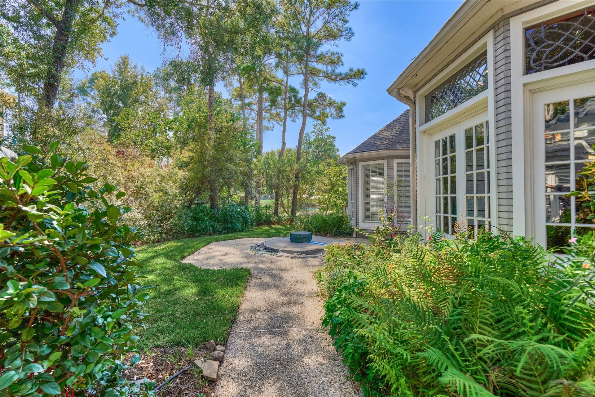 26 S Brokenfern Drive, The Woodlands, TX 77380 This private backyard has an inground spa on an aggregate patio, with a winding aggregate walkway and a lawn framed by mature pines, oaks, and ferns. The spa is reached from the enclosed sunroom and the primary bedroom through French doors with leaded glass transoms, and a bayed wall of the home adds architectural interest. Through the trees, the golf course community stretches out beyond the back of the property.