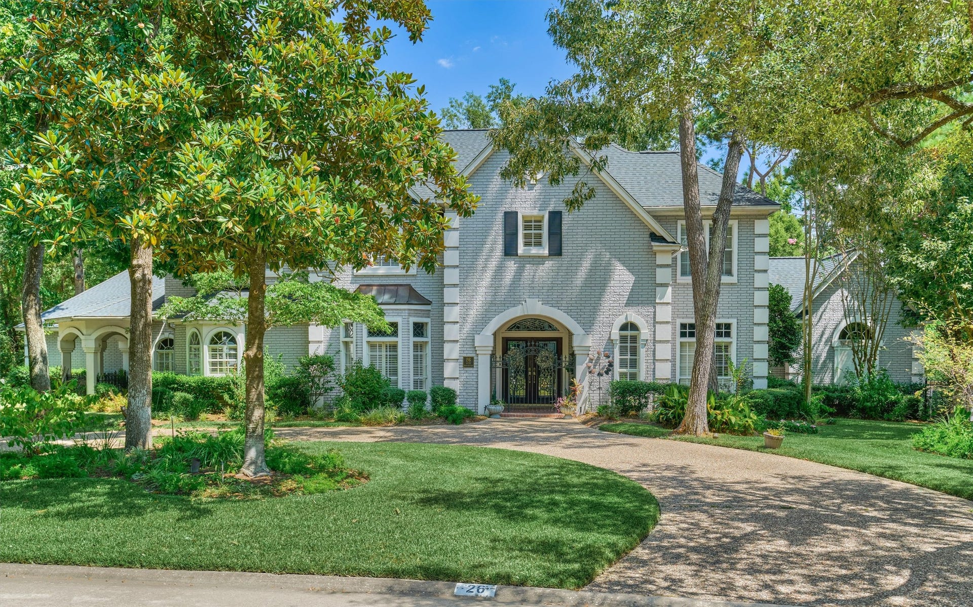 26 S Brokenfern Drive, The Woodlands, TX 77380 Welcome to 26 S Brokenfern. A circular driveway leads to this elegant brick and stucco home, framed by mature magnolia and oak trees. Arched details, an iron-and-glass entry, and lush landscaping give it timeless curb appeal.
