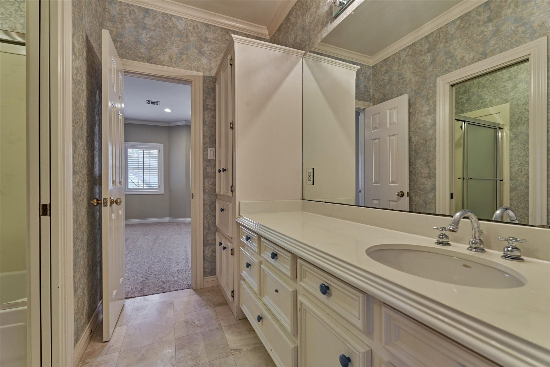 26 S Brokenfern Drive, The Woodlands, TX 77380 This upstairs bath is a Jack and Jill, set between the third bedroom and the game room. A long vanity with raised-panel cabinetry and drawers sits beneath a wall of mirror, with chrome fixtures and a tall built-in linen cabinet for extra storage. A tub and shower combination with a sliding glass door, crown molding, and large-format tile floors complete the room, with a door at each end that opens to the carpeted bedroom and game room.