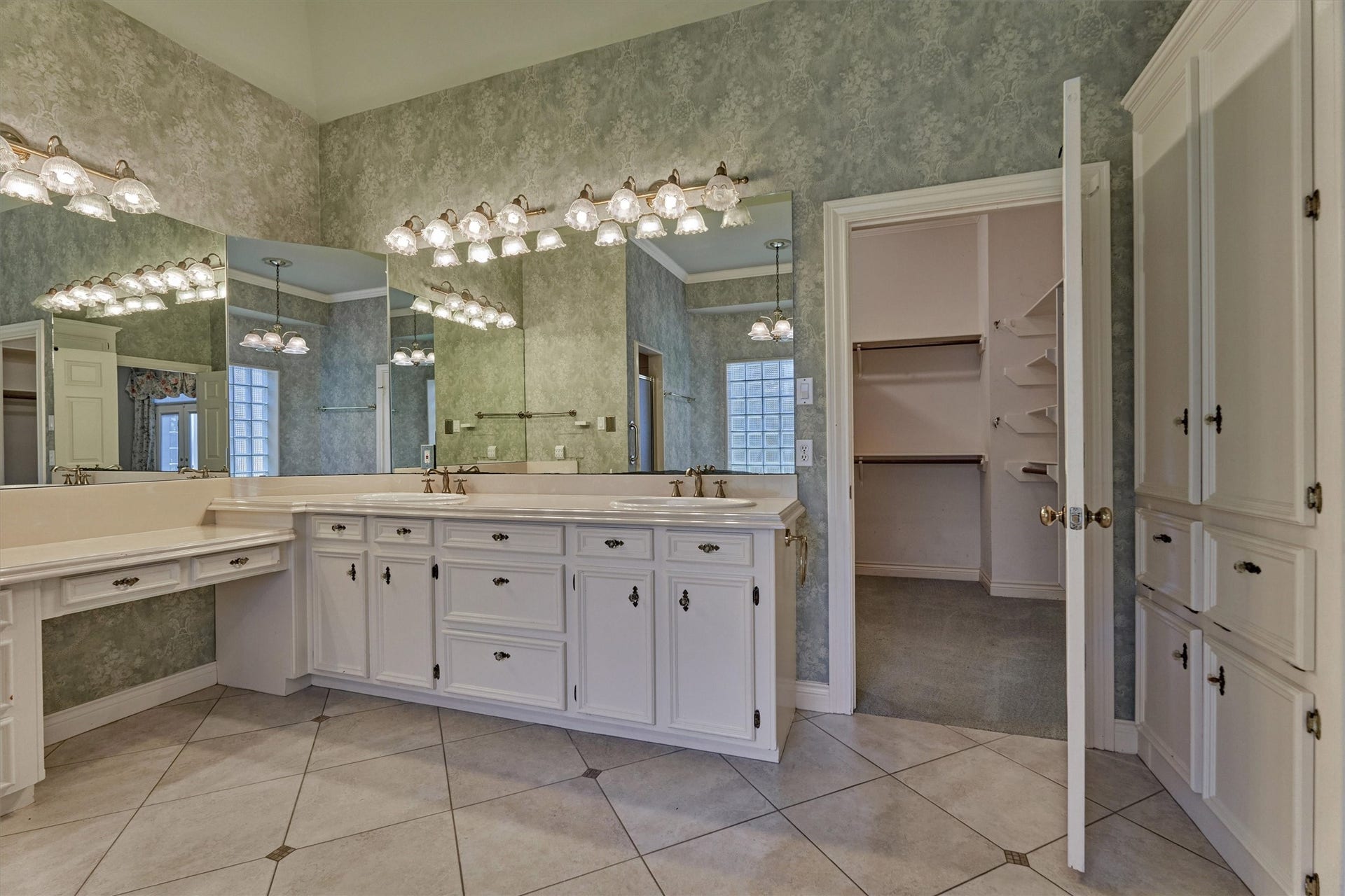 26 S Brokenfern Drive, The Woodlands, TX 77380 Another view of the primary bath shows a double-sink vanity with raised-panel cabinetry and glass-style knobs, a separate makeup vanity with drawers, and a wall of mirrors with multi-light fixtures. A tall built-in linen cabinet adds storage, and the diagonal tile floor has dark accent inserts. A glass block window brings in natural light. The large hers walk-in closet has built-in hanging rods and angled shelving, and his closet is accessed through hers.