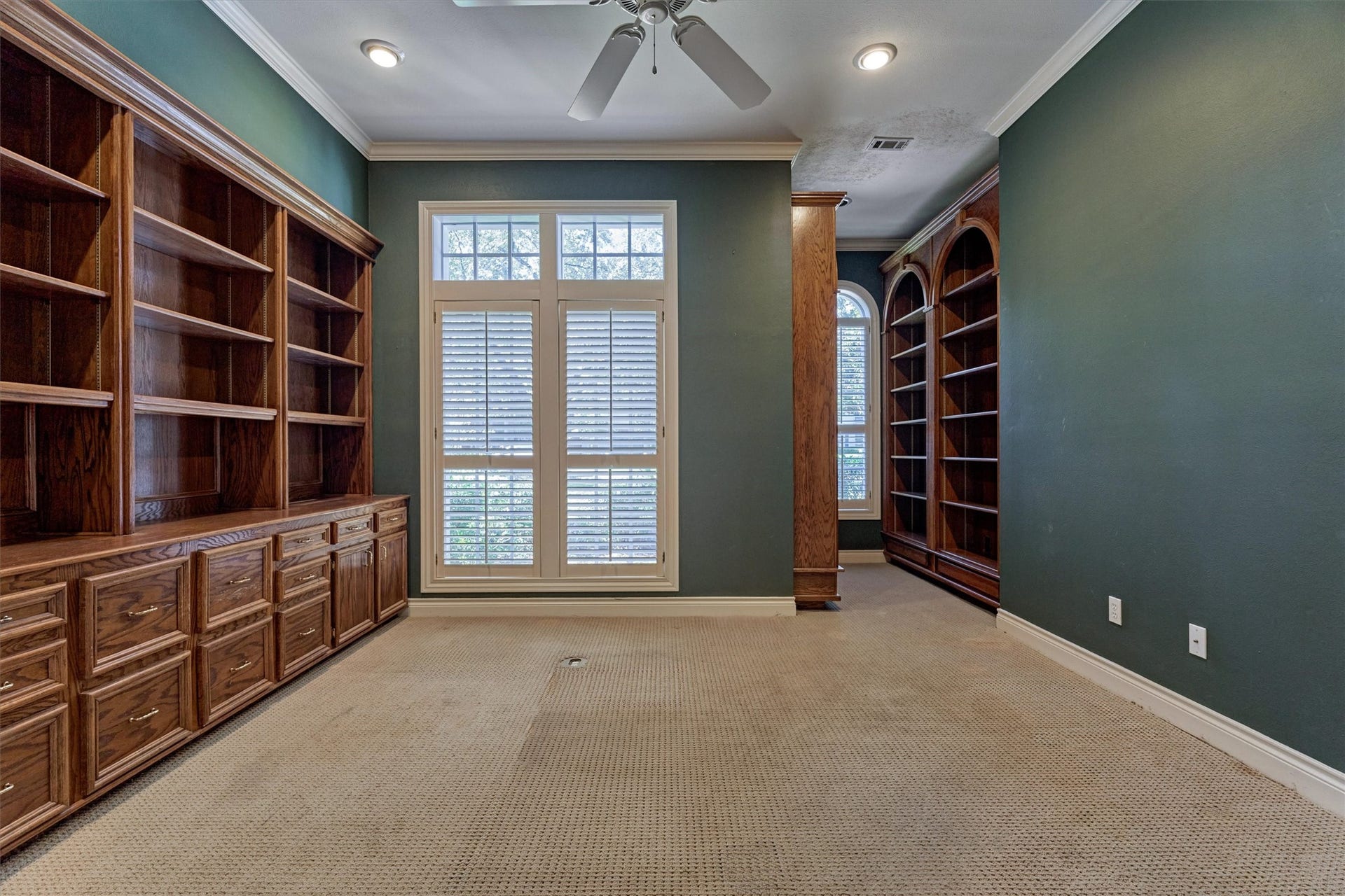 26 S Brokenfern Drive, The Woodlands, TX 77380 This bedroom is currently set up as a private office, with a full wall of oak built-in shelving and cabinetry that has raised-panel drawers, cabinet doors, and a wide work surface. A tall window with a transom and plantation shutters overlooks the front yard. Crown molding, recessed lighting, and a ceiling fan add detail, and a wood-trimmed column separates a second area with arched, floor-to-ceiling bookshelves and an arched window. A walk-in closet completes the room.