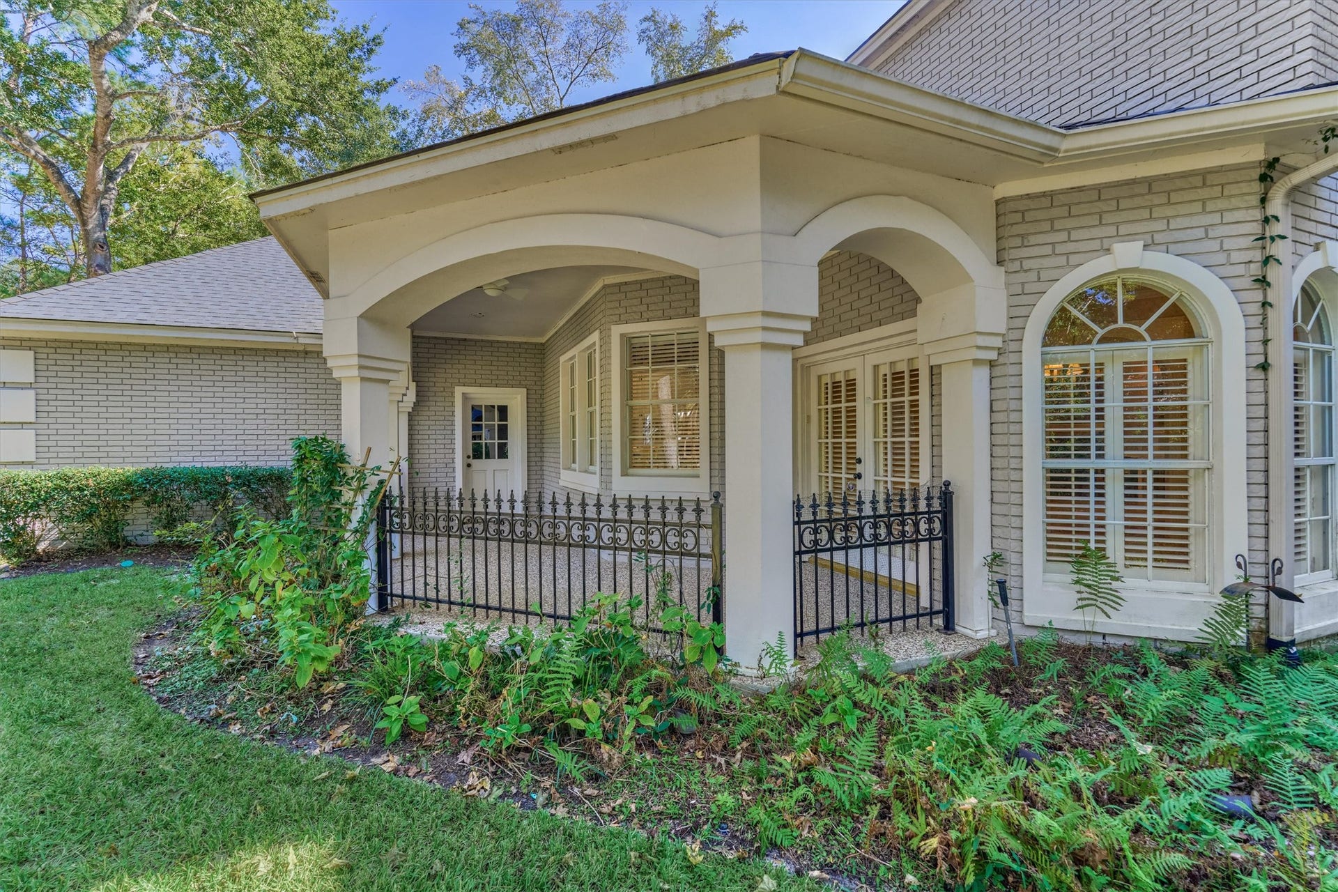26 S Brokenfern Drive, The Woodlands, TX 77380 This covered patio sits off the breakfast room and overlooks the front yard, with direct access to both the breakfast room and the garage. Graceful arches rest on stucco columns, with a ceiling fan overhead and a wrought iron railing with scrollwork and fleur-de-lis finials. French doors with plantation shutters open to the breakfast room, and a separate door at the back of the patio leads to the garage. An aggregate floor, brick walls, and an arched window complete the view.