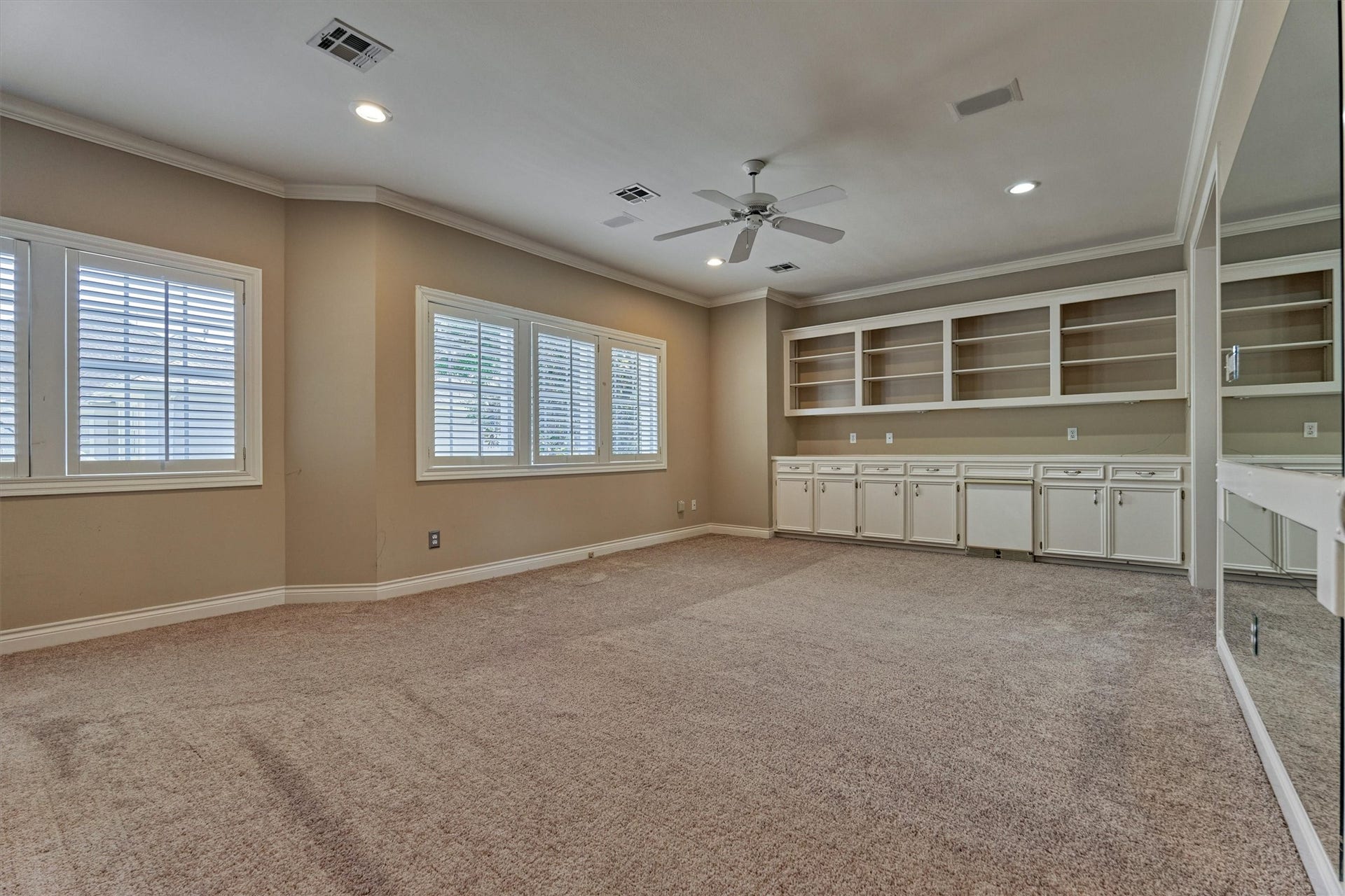 26 S Brokenfern Drive, The Woodlands, TX 77380 This upstairs game room is a large, open space with crown molding, recessed lighting, and a ceiling fan. A full wall of white built-ins has open shelving above and raised-panel cabinets below, with a counter that has room for a mini fridge or beverage center. A wide bank of plantation-shuttered windows lets in plenty of natural light, and a mirrored wall at the right edge adds depth. The Jack and Jill bath is steps away, and plush carpet runs throughout.