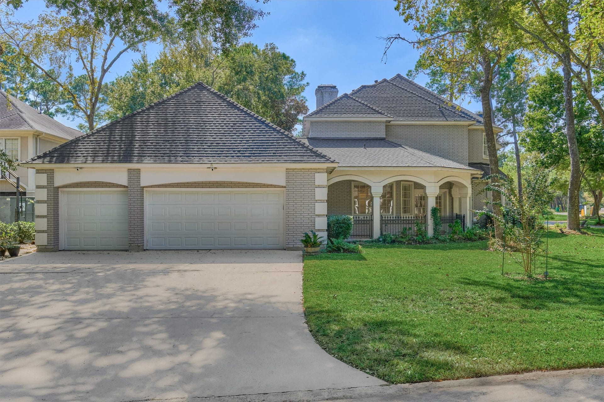 26 S Brokenfern Drive, The Woodlands, TX 77380 Side view of this corner-lot beauty, featuring a three-car garage with a wide driveway for plenty of parking. The arched, covered front patio is a welcoming spot for morning coffee or evening relaxation, all surrounded by a lush lawn and mature trees.