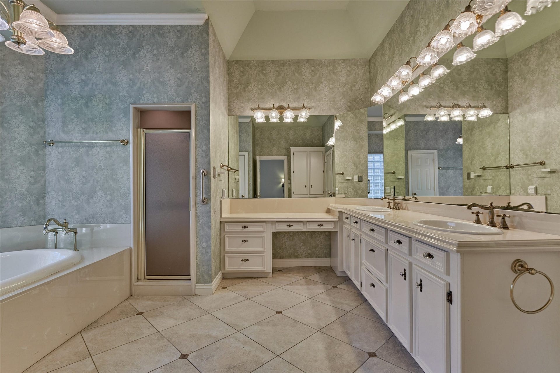 26 S Brokenfern Drive, The Woodlands, TX 77380 This primary bath has a vaulted ceiling and a diagonal-set tile floor with dark accent inserts. A double-sink vanity with raised-panel cabinetry, crystal-style knobs, and bronze-finish faucets sits beneath a wall of mirrors, and a separate makeup vanity with drawers adds a seated dressing area. A drop-in soaking tub with a vintage-style deck faucet and a separate walk-in shower complete the room, along with a private water closet and a glass block window.