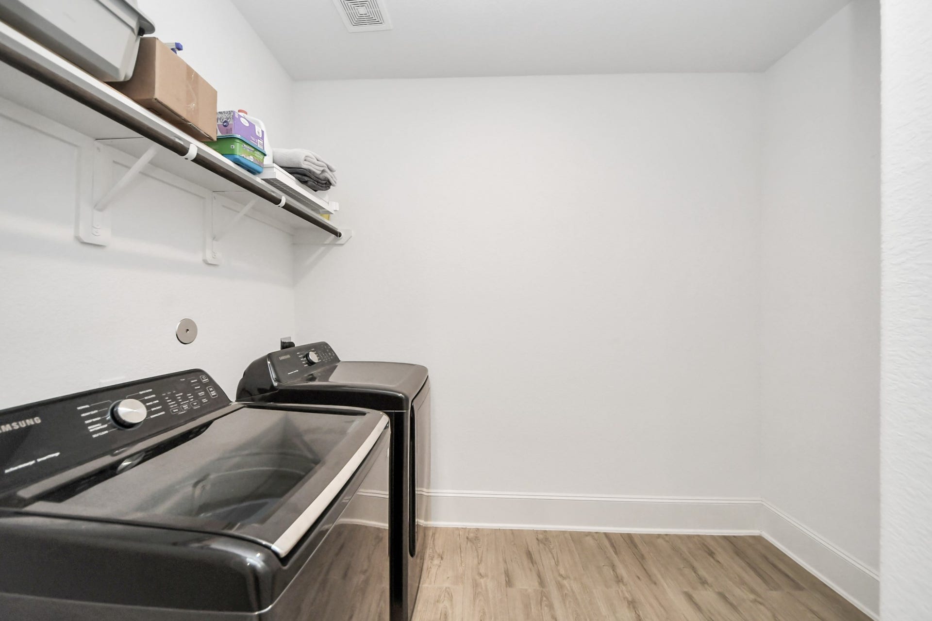 Spacious laundry room with shelving, and sleek wood look tile flooring. Oversized and recently purchased washer and dryer are available for purchase.