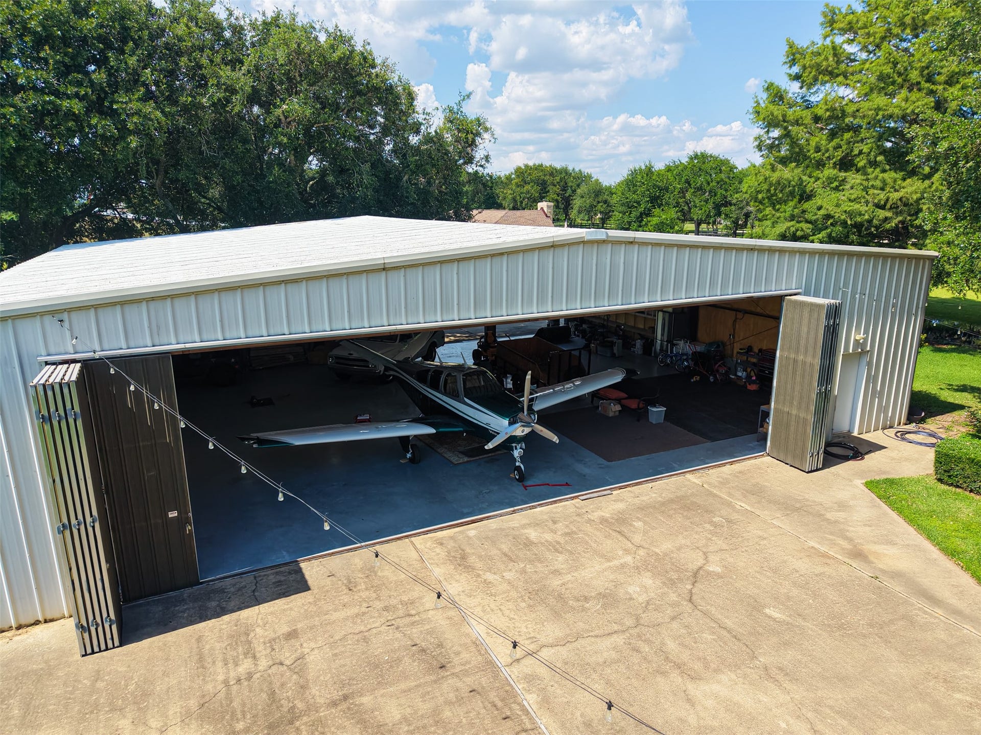 27 E Kitty Hawk Street, Richmond, TX 77406 This photo features a spacious airplane hangar with wide-open doors, housing a aircraft. The hangar is well-lit with ample space for storage and maintenance equipment, set on a paved surface with surrounding greenery. Ideal for aviation enthusiasts.