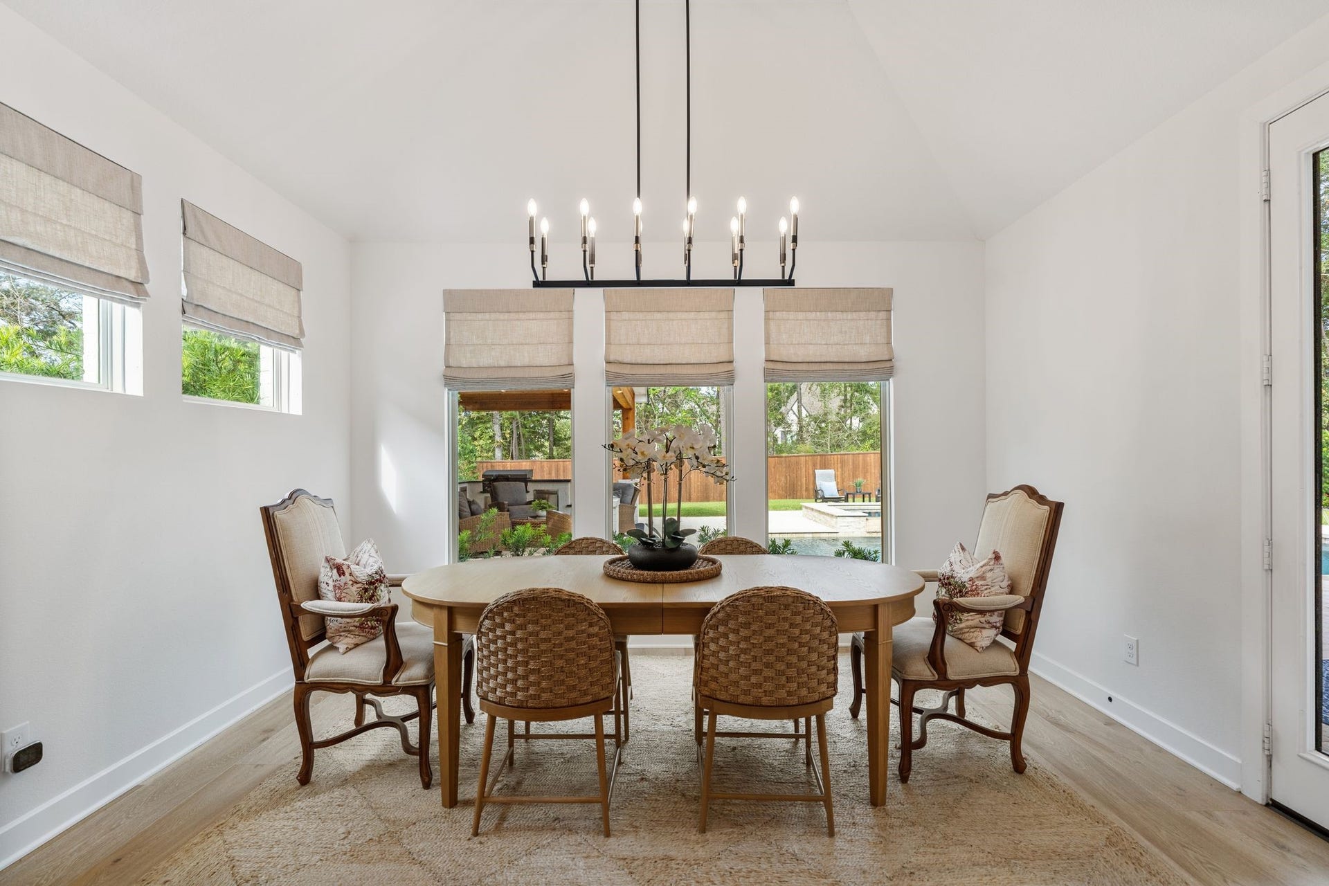 Dining area featuring a stunning designer chandelier, 9" white oak flooring, custom Roman shades, and walls of windows showcasing picturesque views of the private backyard oasis.
