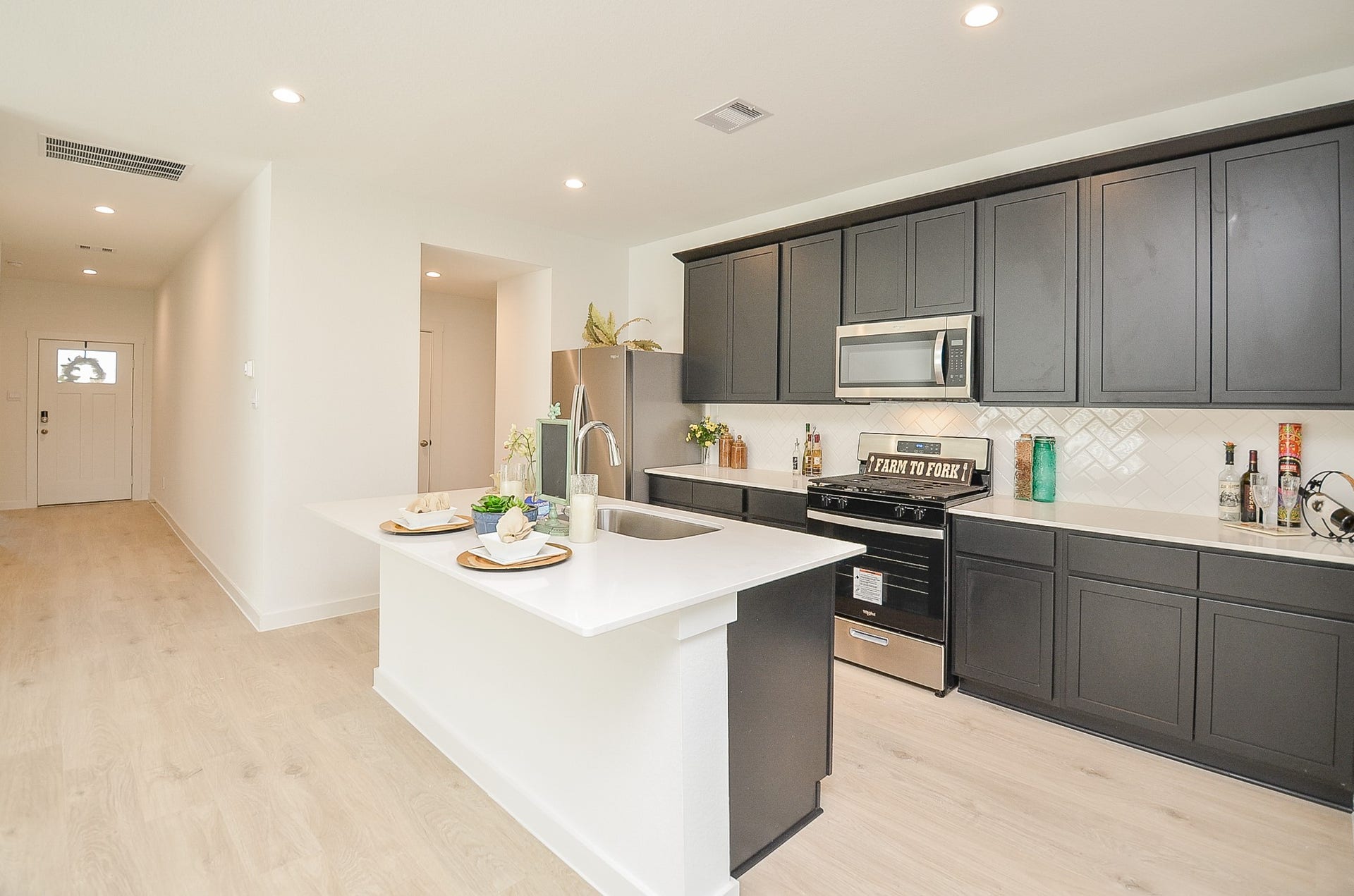 7814 Bel Fiore Drive, Richmond, TX 77406 A modern kitchen featuring sleek dark cabinetry, stainless steel appliances, and a spacious island with bar seating, seamlessly connected to a bright hallway.
