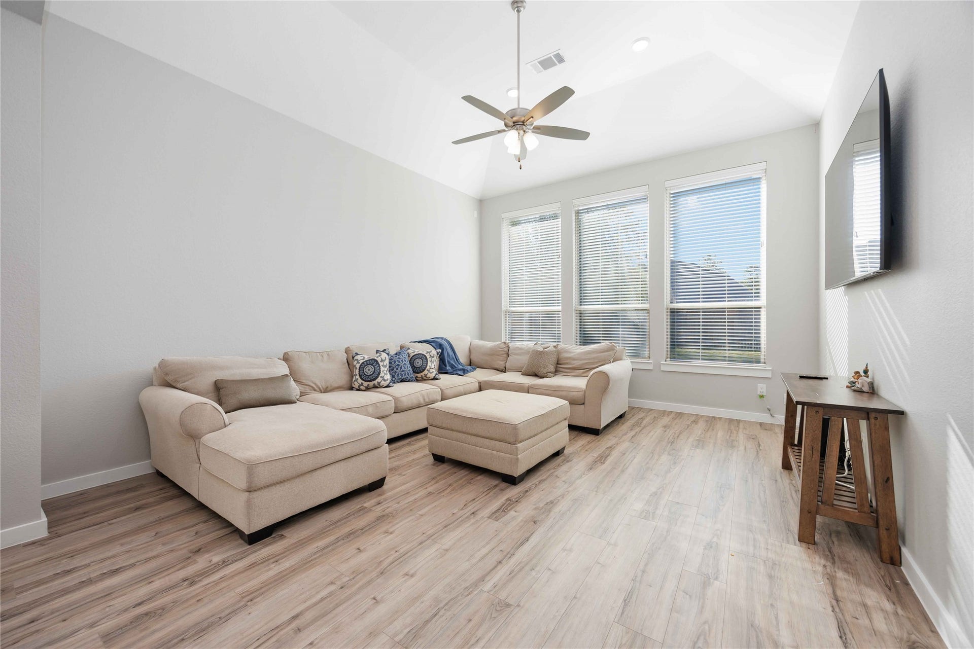 12610 Woodbourne Forest Drive, Humble, TX 77346 Bright and spacious living room with large windows, wood flooring, and a neutral-toned sectional sofa. Features high ceilings, a ceiling fan, and a wall-mounted TV for a modern touch.