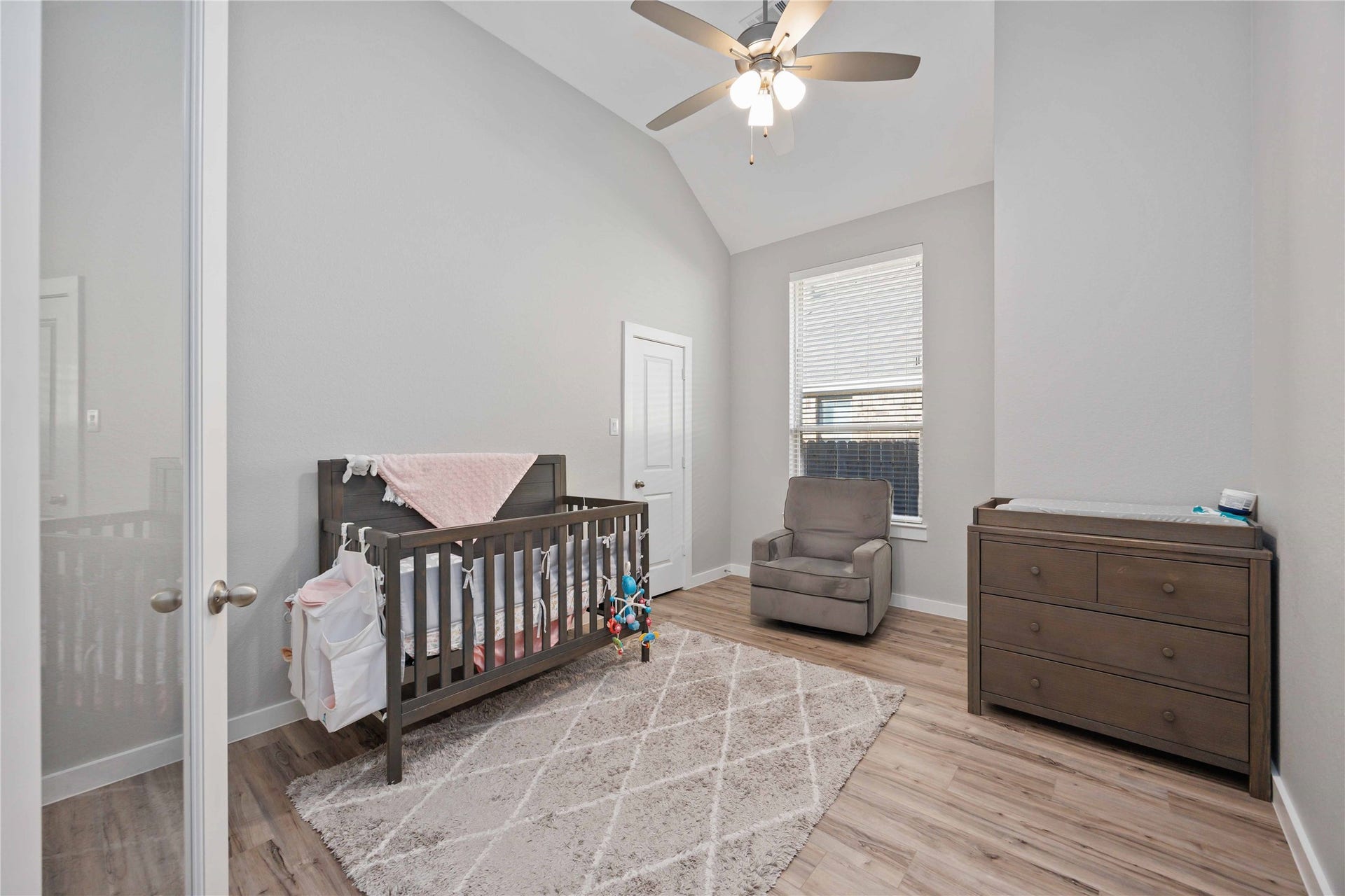 12610 Woodbourne Forest Drive, Humble, TX 77346 This cozy nursery features a crib, a comfortable armchair, and a dresser, all set against soft gray walls and wood flooring. A ceiling fan and large window provide ample light and ventilation.