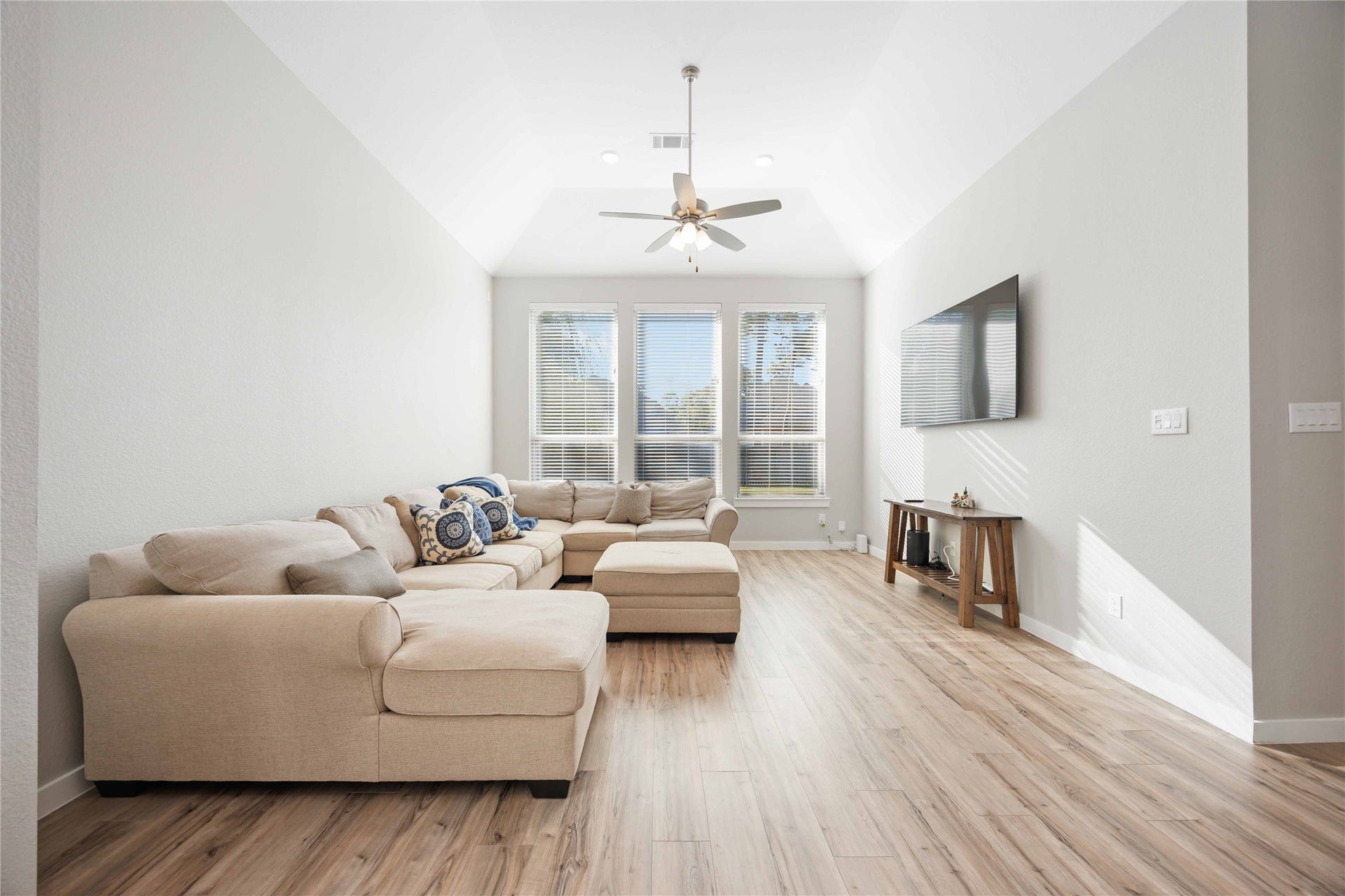 12610 Woodbourne Forest Drive, Humble, TX 77346 Bright and spacious living room with high ceilings, featuring a cozy beige sectional sofa, wood flooring, and a wall-mounted TV. Large windows let in plenty of natural light.