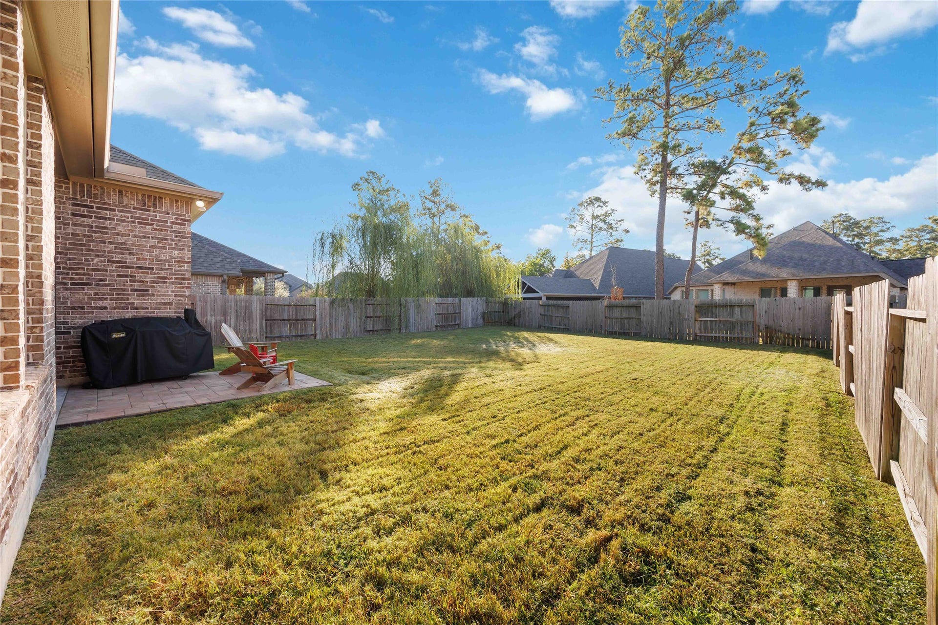 12610 Woodbourne Forest Drive, Humble, TX 77346 This photo shows a spacious backyard with a well-maintained lawn, bordered by a wooden fence. There's a small patio area with outdoor seating and a grill, perfect for relaxation or entertaining. The yard is surrounded by other houses, offering privacy and a serene atmosphere.