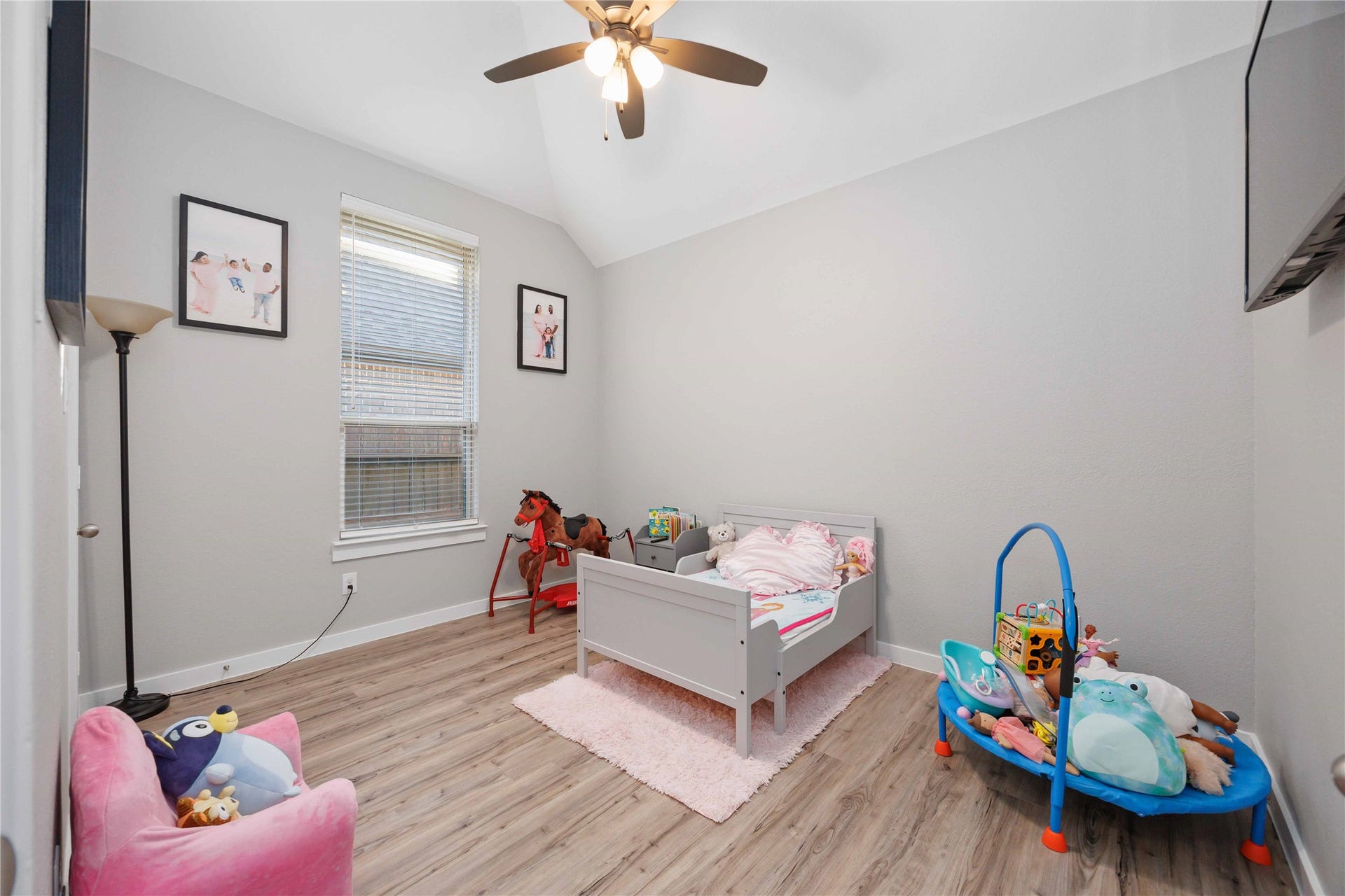 12610 Woodbourne Forest Drive, Humble, TX 77346 This cozy child's bedroom features a light gray color scheme with wood-look flooring. It includes a small bed, plush toys, a floor lamp, and wall art. The room is well-lit with natural light from a window, complemented by a ceiling fan with lights.