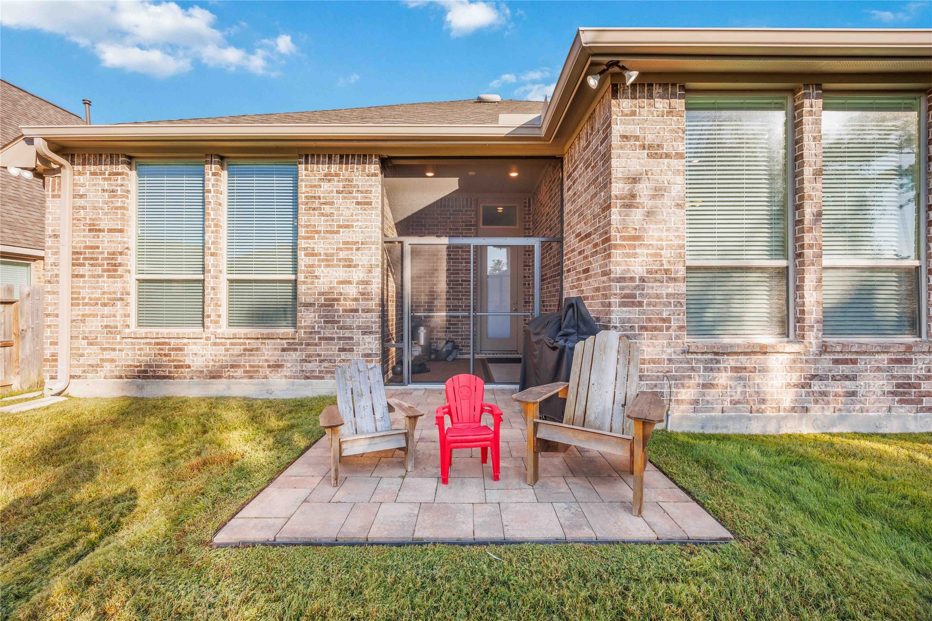 12610 Woodbourne Forest Drive, Humble, TX 77346 Charming brick patio with two wooden chairs and a child's chair, perfect for relaxing. The backyard features a well-maintained lawn and a covered entryway, ideal for outdoor enjoyment.