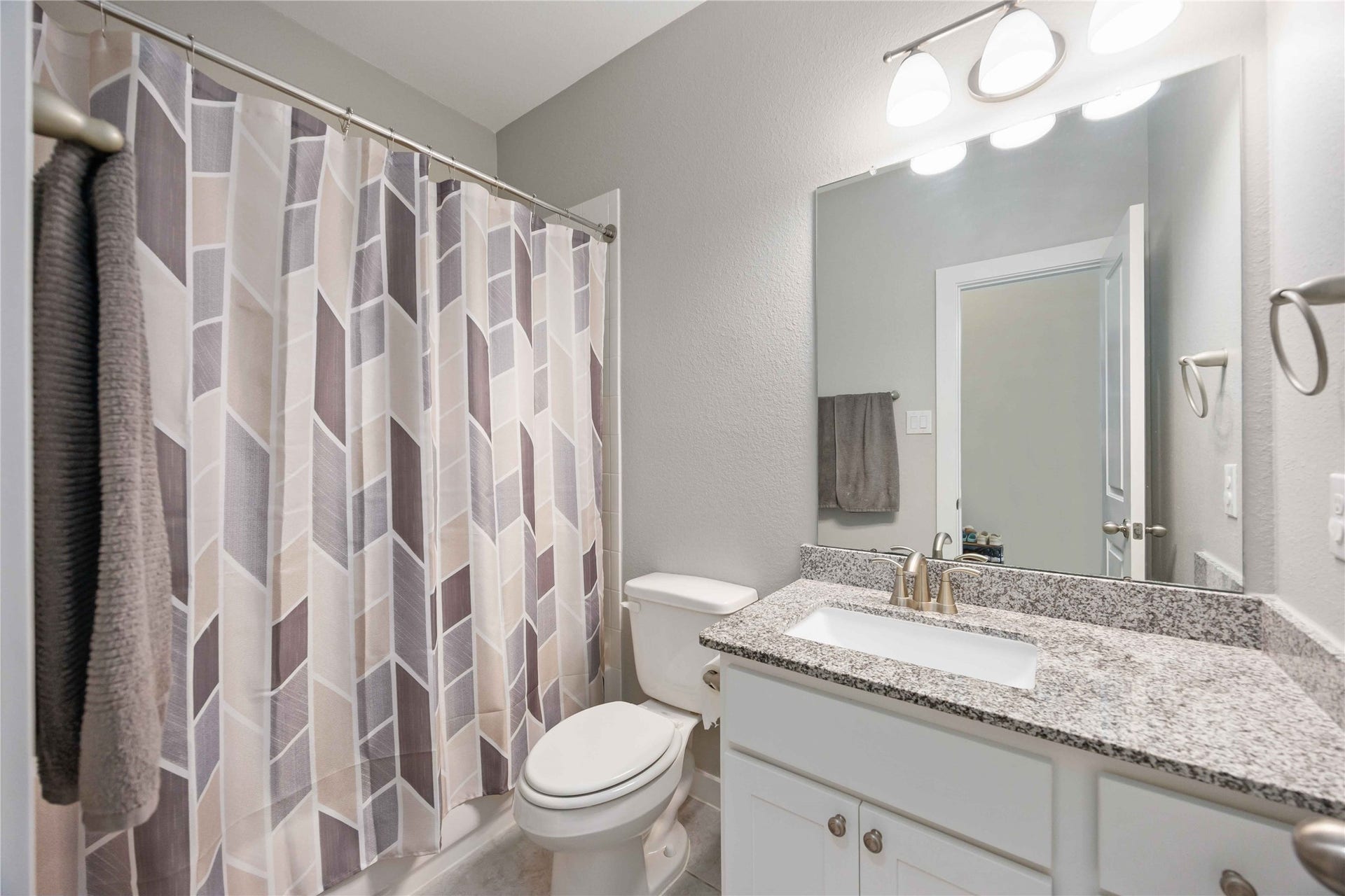12610 Woodbourne Forest Drive, Humble, TX 77346 This bathroom features a modern design with a sleek granite countertop, white cabinetry, and a well-lit mirror. It includes a shower with a stylish geometric-patterned curtain, and neutral wall tones create a clean, inviting atmosphere.