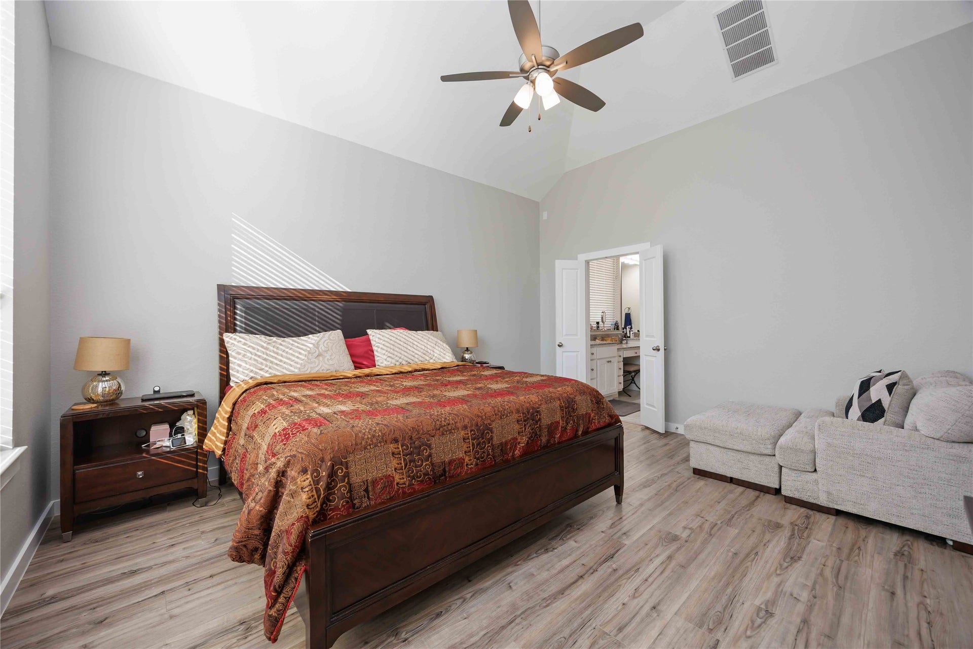 12610 Woodbourne Forest Drive, Humble, TX 77346 Spacious bedroom with high ceilings, featuring a large bed with elegant bedding, a comfy corner sofa, and an en-suite bathroom. The room has a modern ceiling fan, neutral walls, and plenty of natural light from a nearby window.