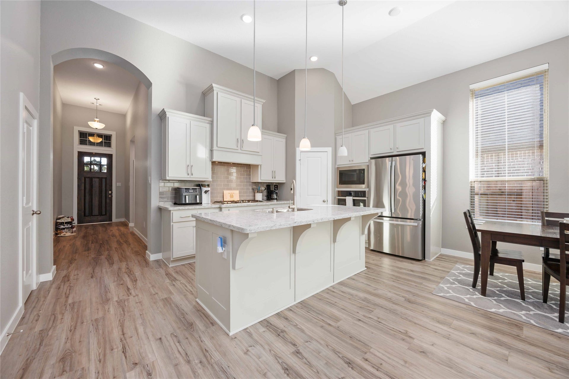 12610 Woodbourne Forest Drive, Humble, TX 77346 This modern kitchen features high ceilings, sleek stainless steel appliances, and a spacious island with pendant lighting. The open layout includes a dining area next to large windows for natural light. The wood-style flooring adds warmth, and the entryway access provides a welcoming flow.