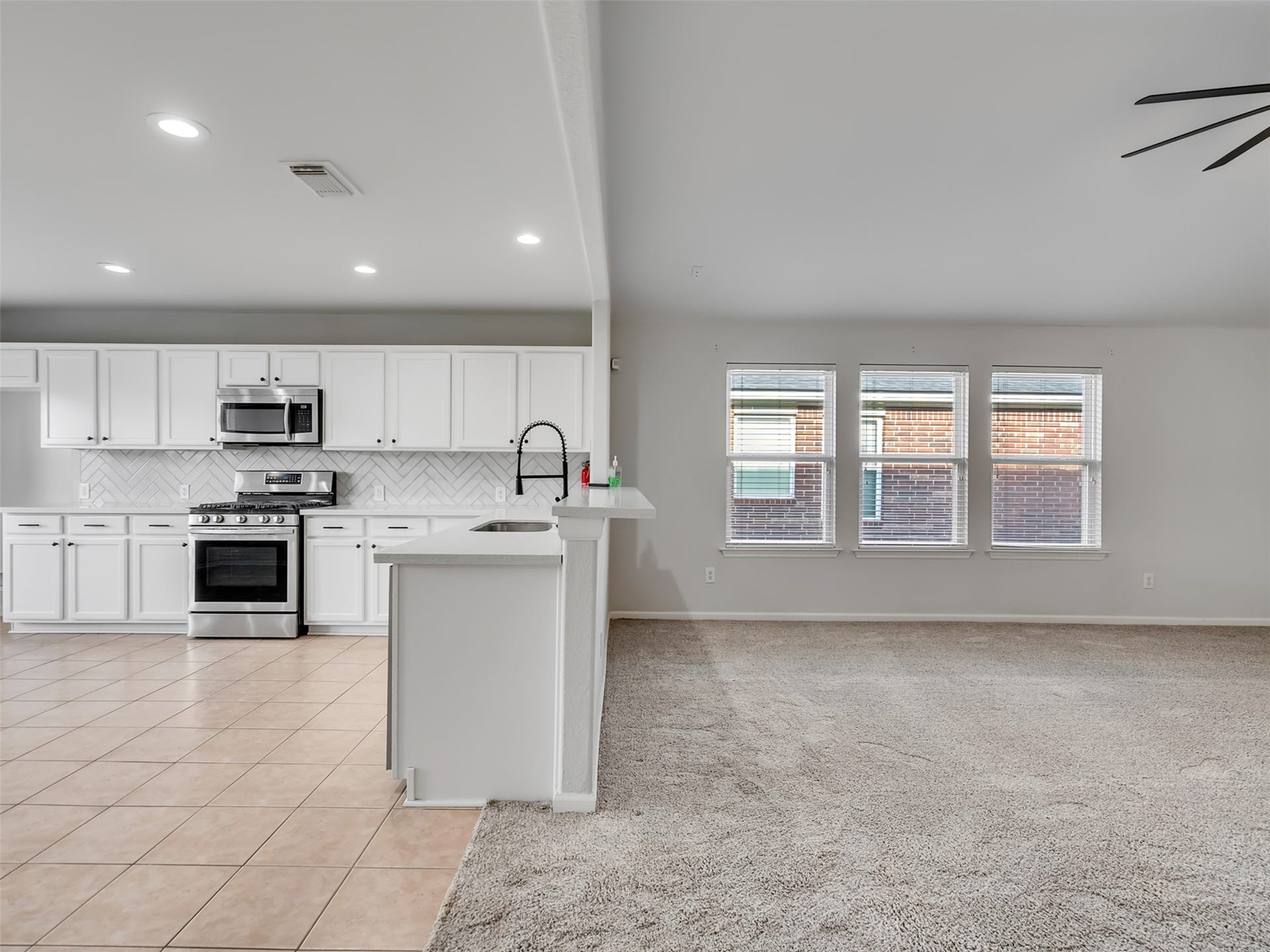 21518 Gannet Peak Way, Katy, TX 77449 A spacious, open-concept living area featuring a modern kitchen with white cabinetry, stainless steel appliances, and a stylish backsplash. The room is well-lit by natural light from multiple windows and has a combination of tiled and carpeted flooring.