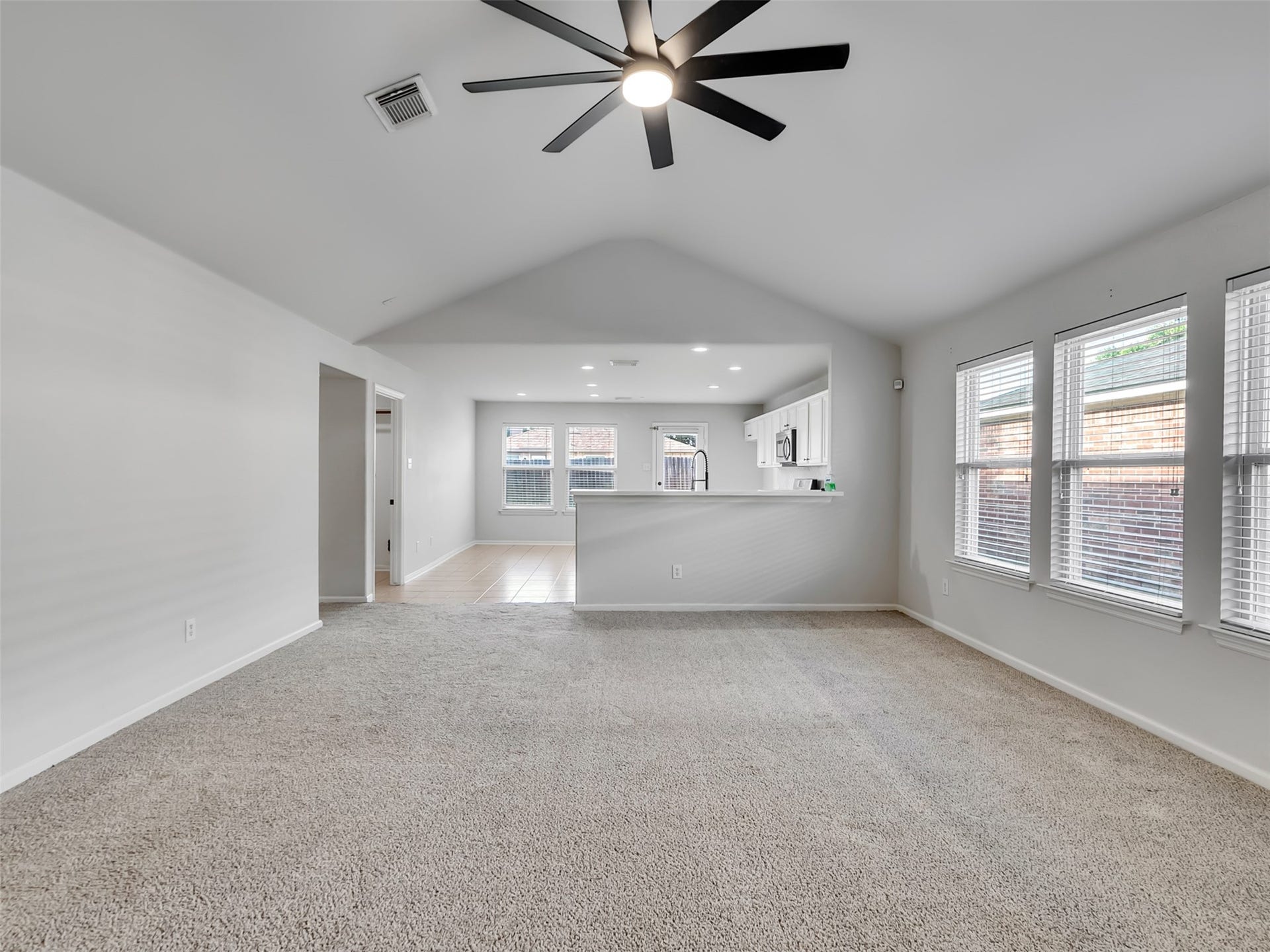 21518 Gannet Peak Way, Katy, TX 77449 Spacious living area with neutral carpet, high ceilings, and a modern ceiling fan. The room is brightened by large windows and opens into a kitchen with white cabinetry and a breakfast bar.